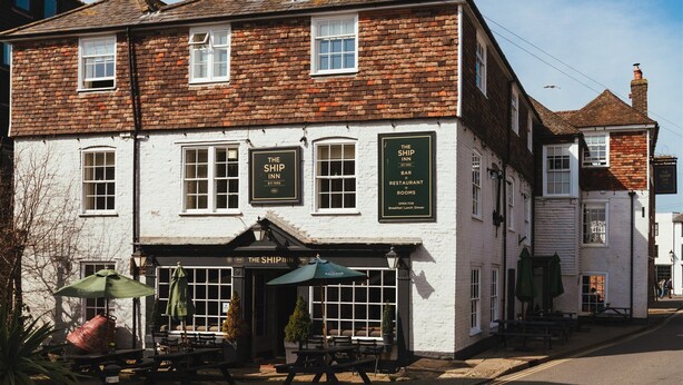 A traditional brick building with a white facade, featuring a pub named "The Ship Inn" with outdoor seating and umbrellas.