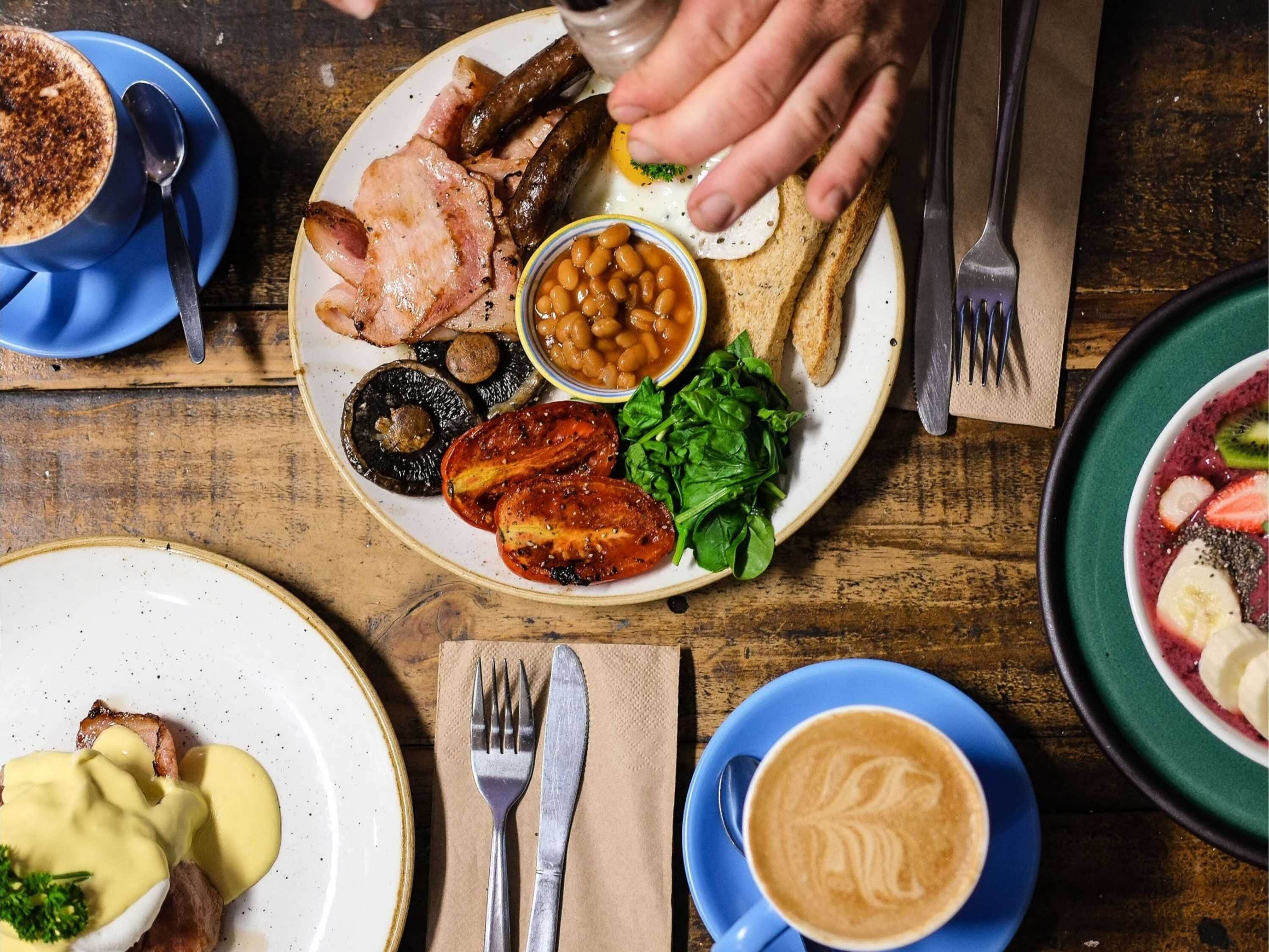 Ein Frühstückstisch mit Eiern, Speck, Pilzen, Bohnen, Tomaten und Grünzeug, dazu eine Tasse Kaffee und eine Schüssel Suppe.