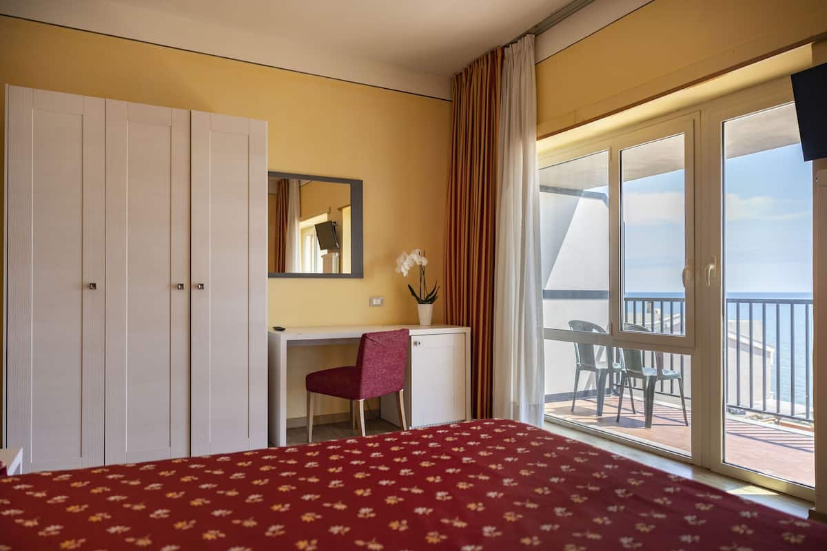 Une chambre d’hôtel comprenant un lit, un bureau, une chaise, une armoire, une plante en pot et un balcon avec vue.