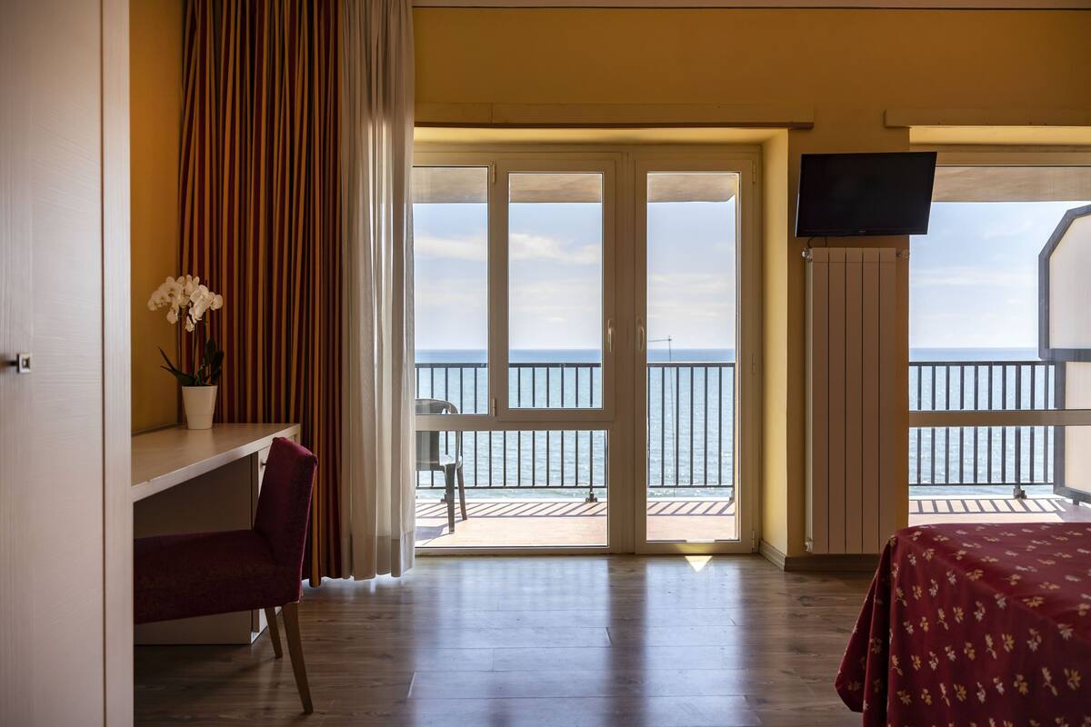Une chambre d’hôtel avec un plancher en bois, un bureau avec une chaise, un lit recouvert d’une courtepointe à motifs floraux, un téléviseur fixé au mur et un balcon avec vue sur la mer.