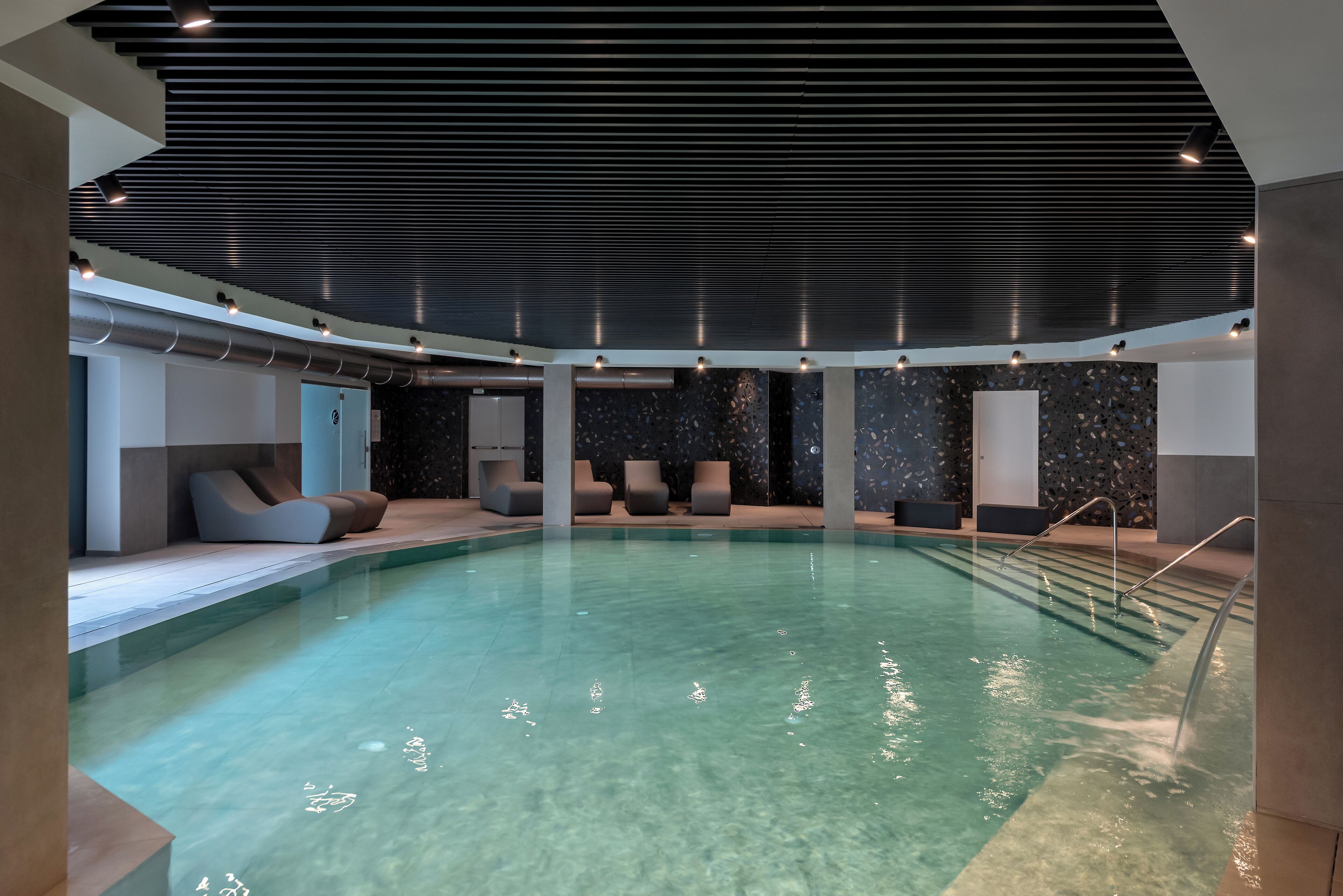 An indoor swimming pool with clear water, modern seating, and a dark ceiling.