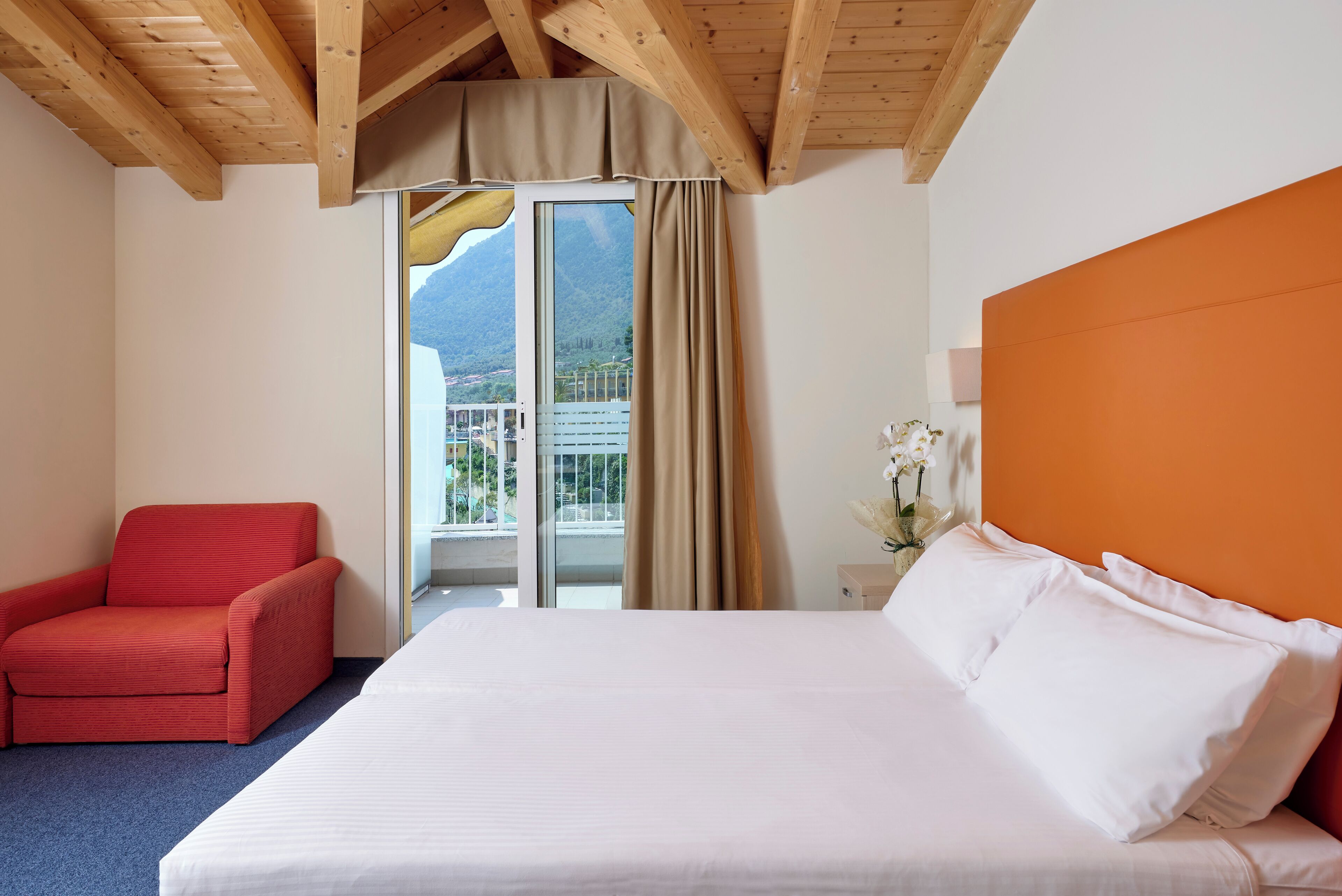 Ein Hotelzimmer mit einem Bett, einem roten Sessel, einem Fenster mit Bergblick und einer Holzbalkendecke.