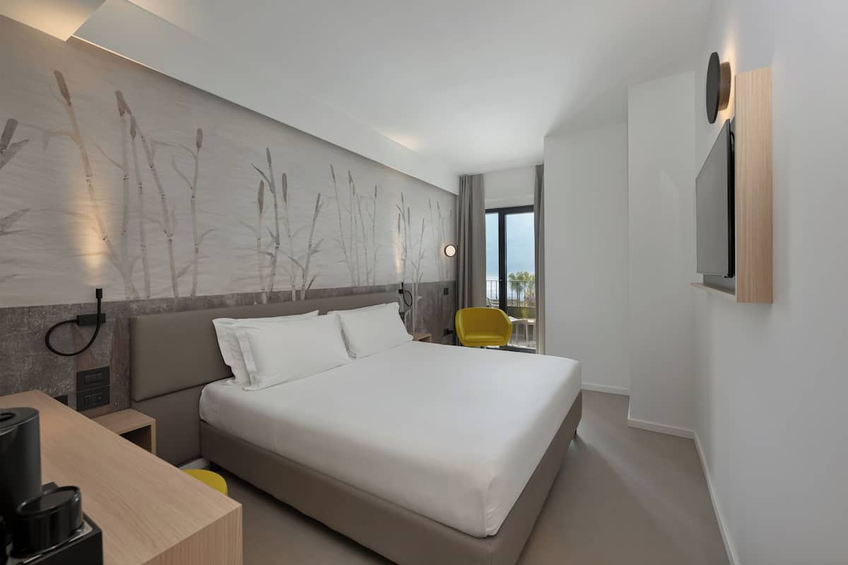 Une chambre d’hôtel moderne dotée d’un grand lit, d’un téléviseur à écran plat fixé au mur, d’un petit bureau avec une lampe et offrant une vue sur l’extérieur.