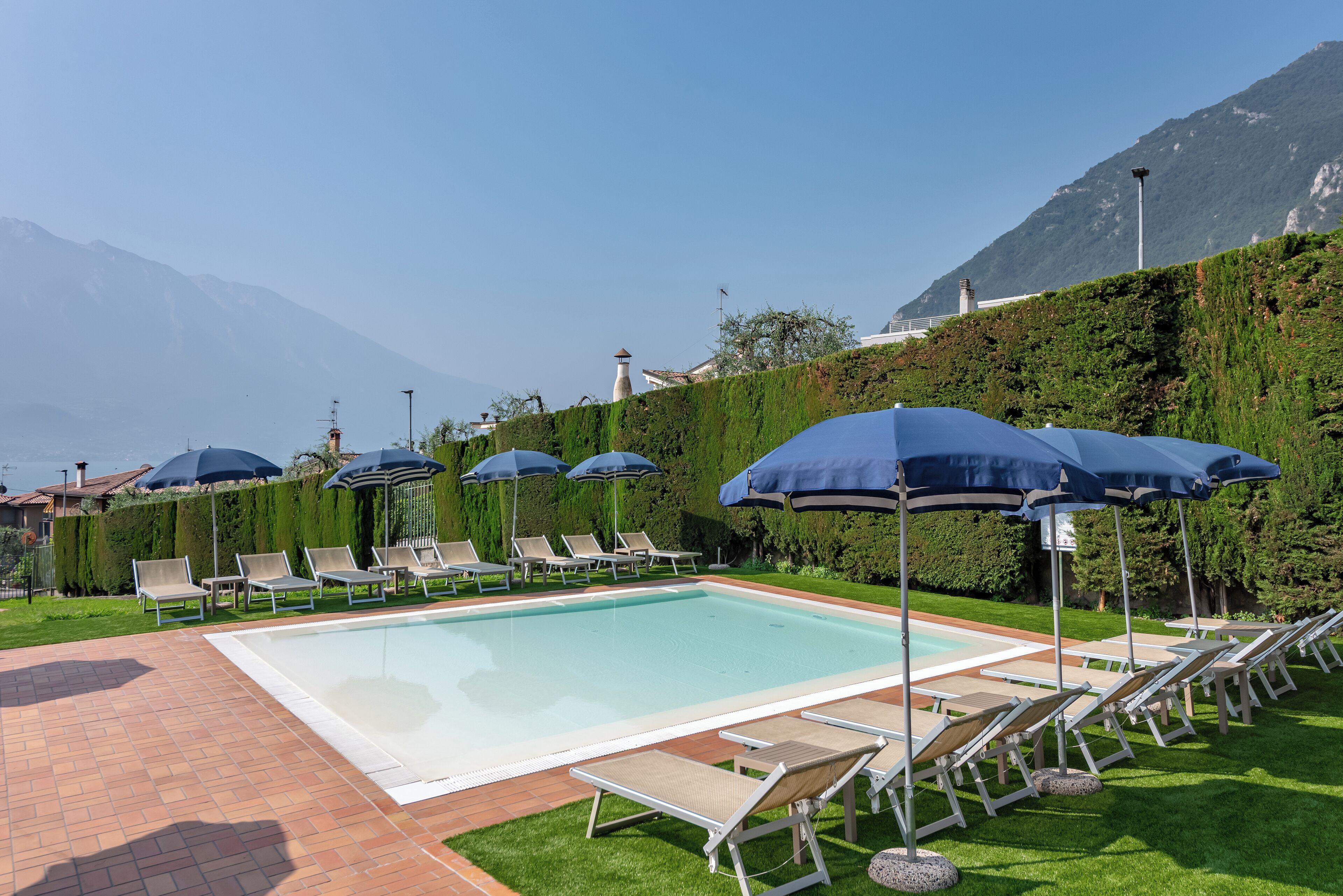 Piscina con sillones de descanso y sombrillas, rodeada por un seto cuidado y con montañas al fondo.