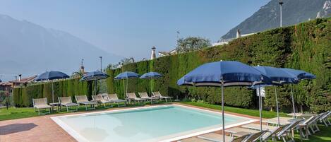 A swimming pool with lounge chairs and umbrellas, surrounded by a well-maintained hedge and a mountain backdrop.
