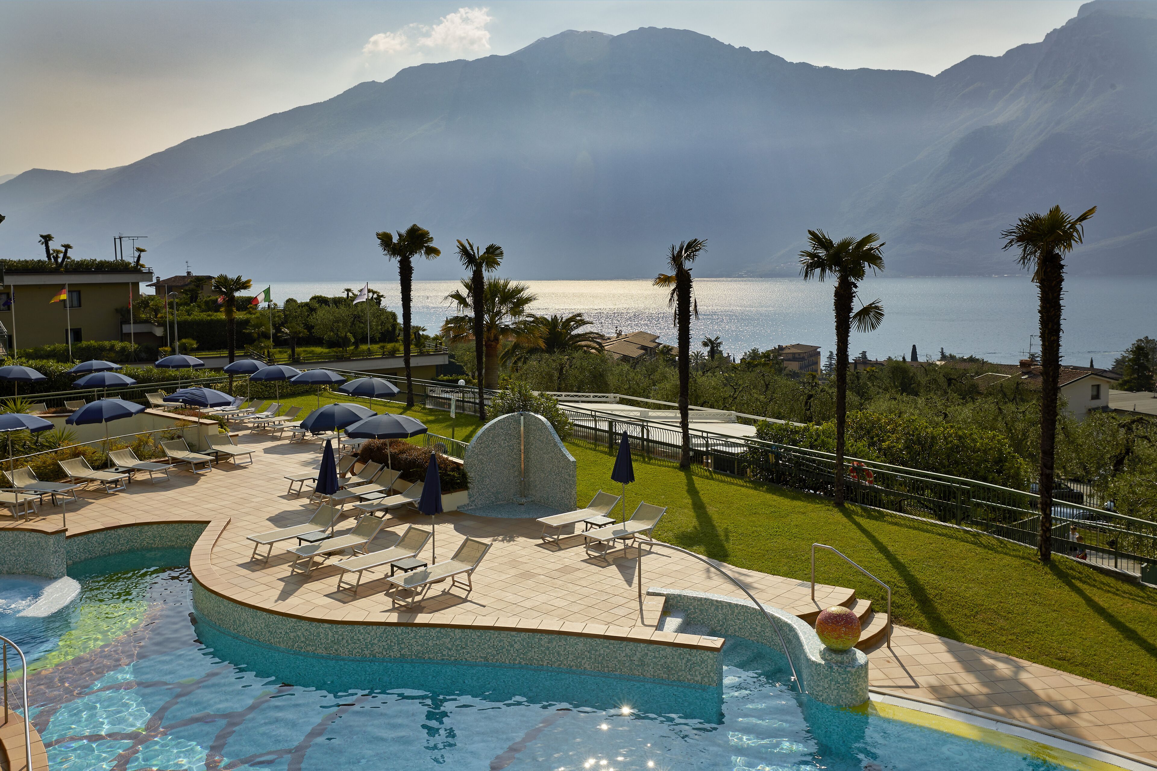 A pool area with lounge chairs and umbrellas, surrounded by palm trees and a view of a lake and mountains.