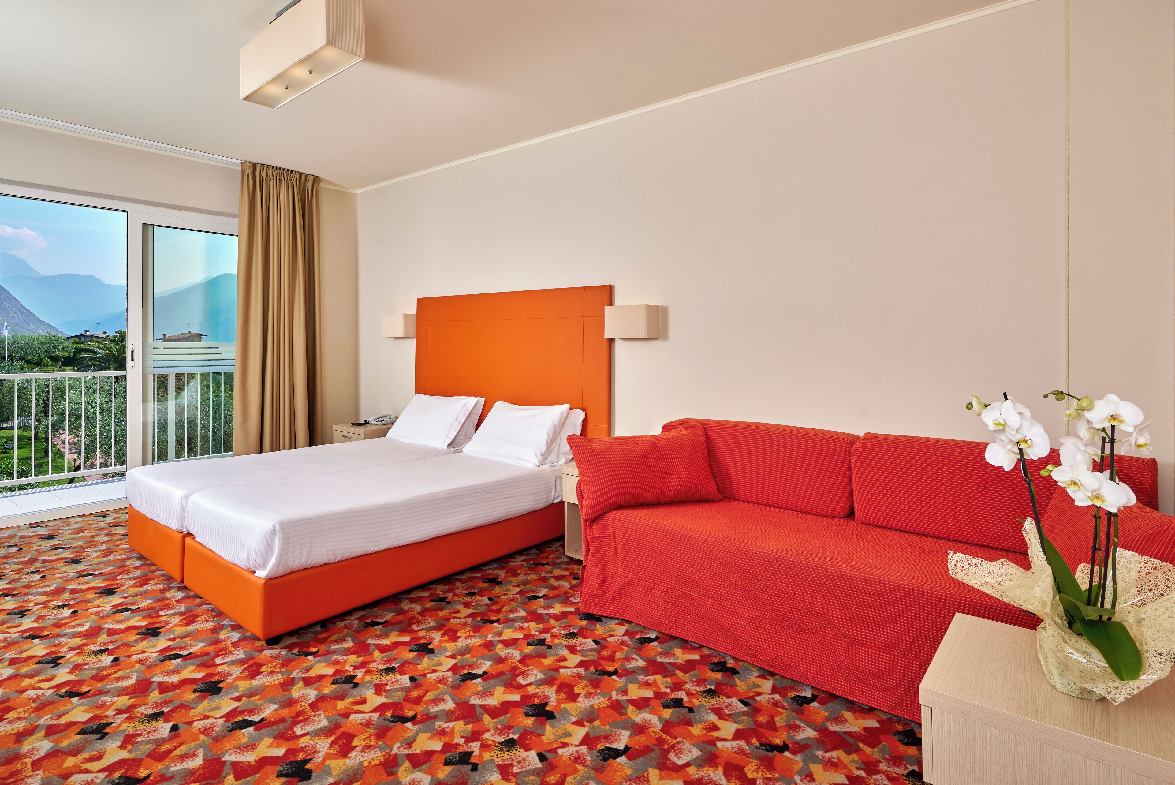 A hotel room with a bed, a red sofa, a nightstand, and a vase of white orchids.