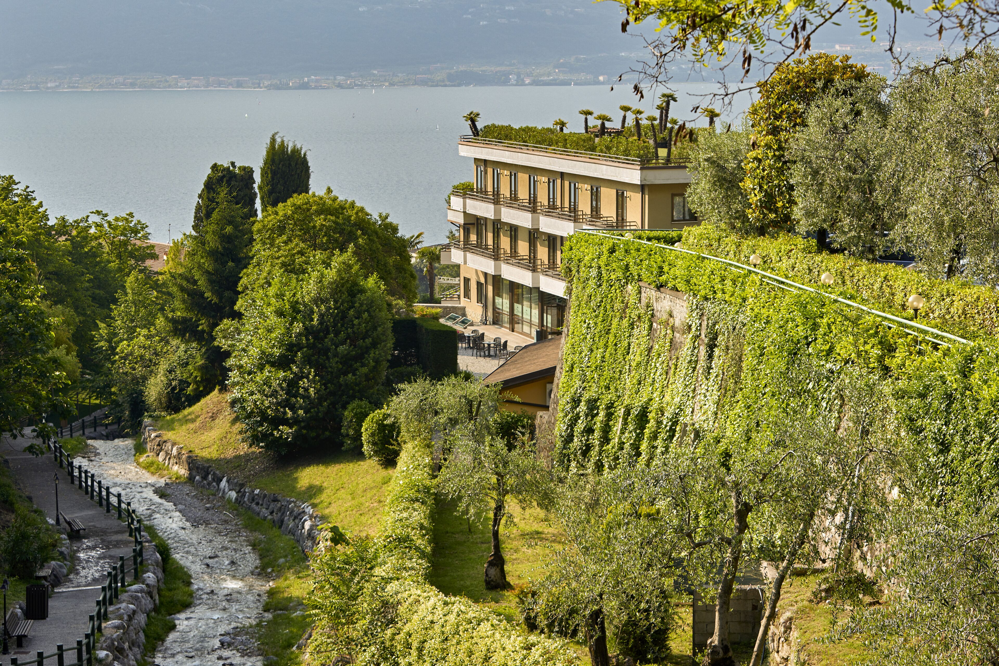 Un edificio moderno con un balcone affacciato su un fiume, circondato da vegetazione lussureggiante e un vialetto in pietra.