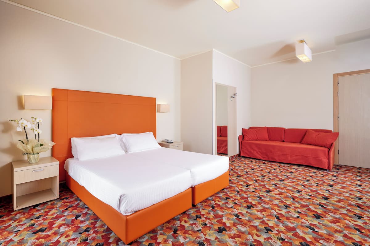Une chambre d’hôtel avec un grand lit, une table de chevet, un canapé rouge et une moquette à motifs.