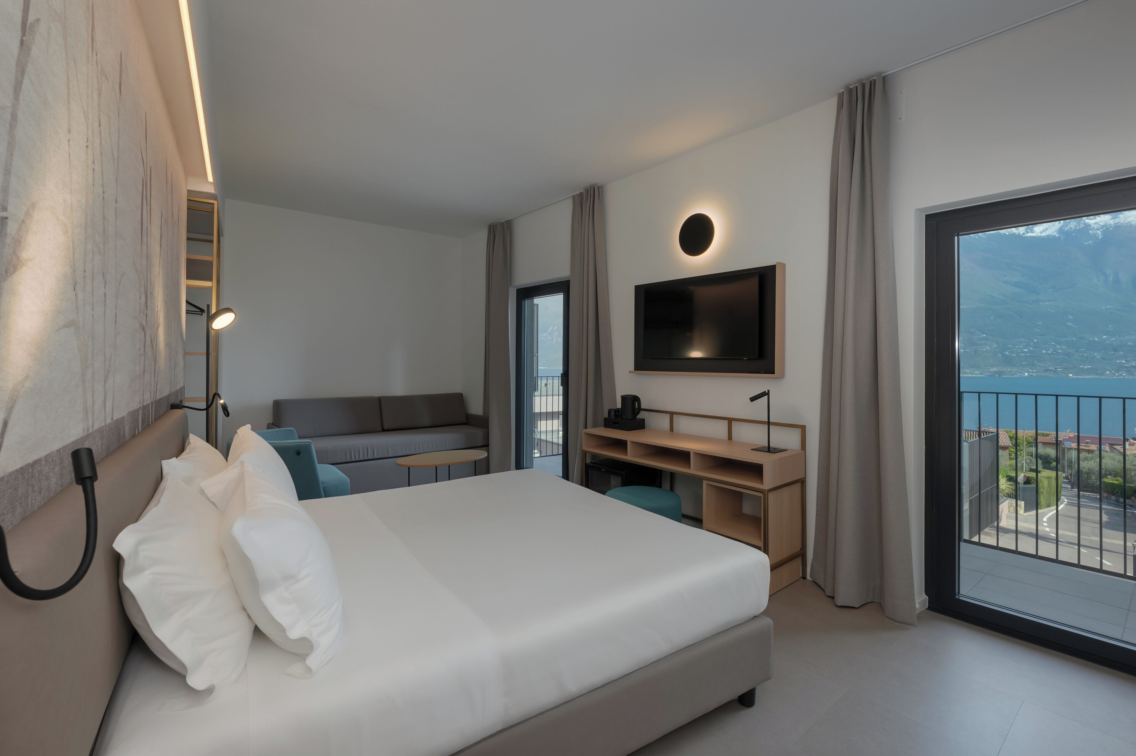 A modern hotel room with a large bed, a TV mounted on the wall, a desk, and a balcony with a view.