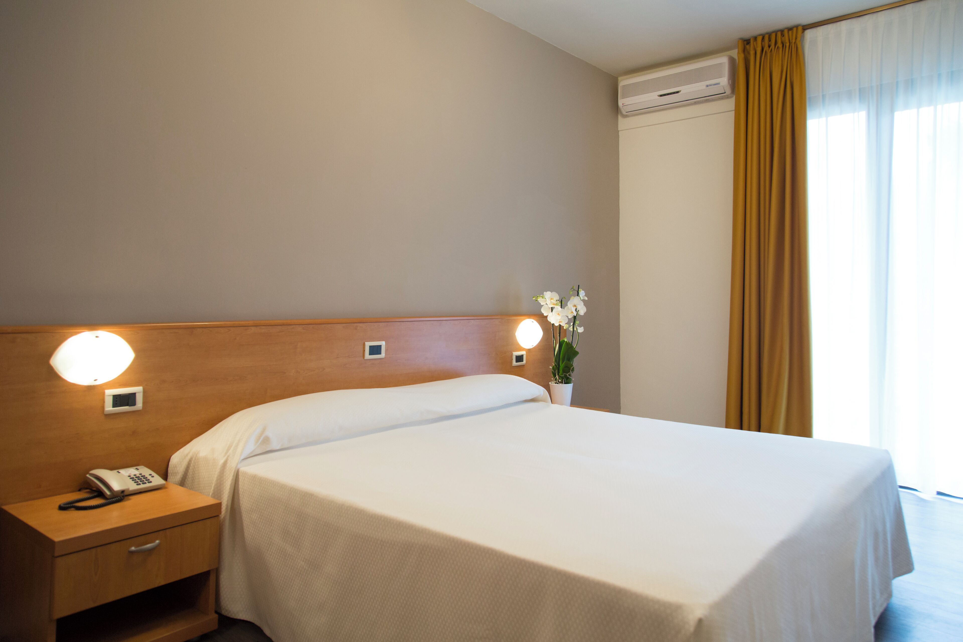 A hotel room with a neatly made bed, bedside lamps, a telephone, and a potted plant.