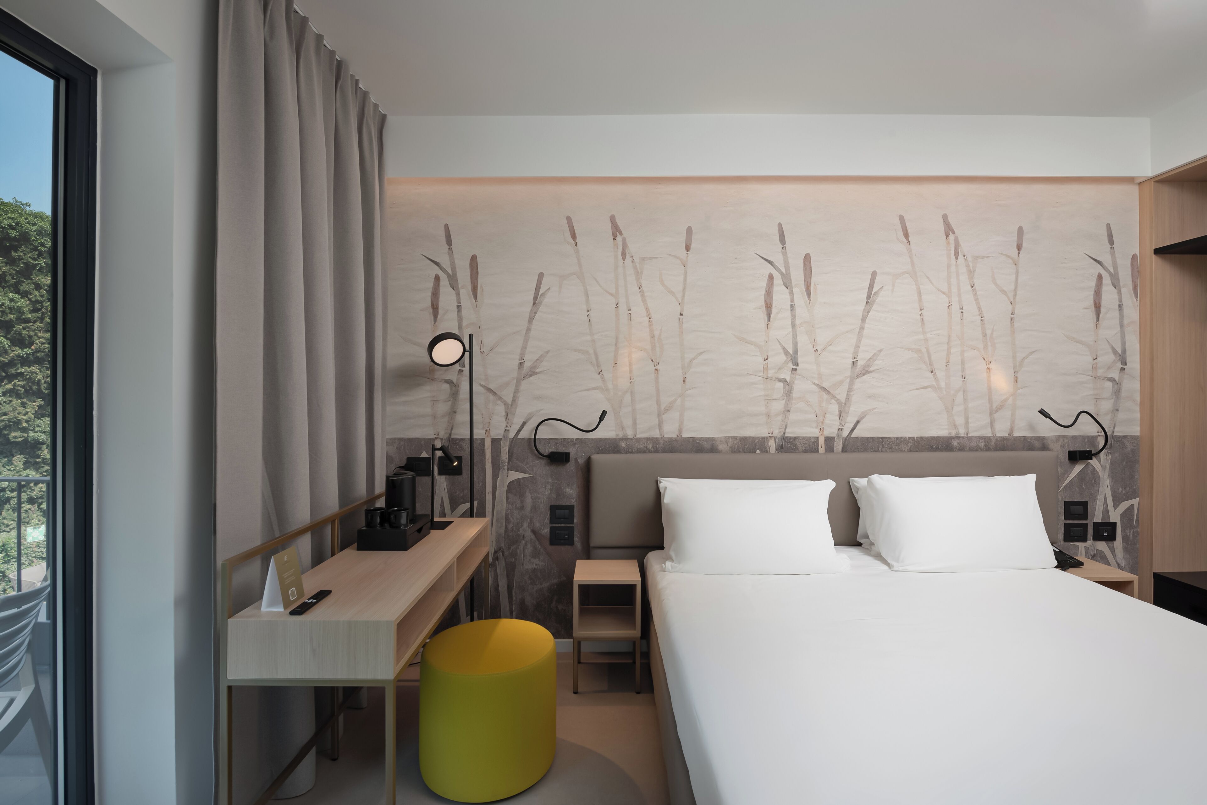 Camera d'albergo moderna con un letto grande, una scrivania, una sedia, un tavolino e un poggiapiedi giallo.