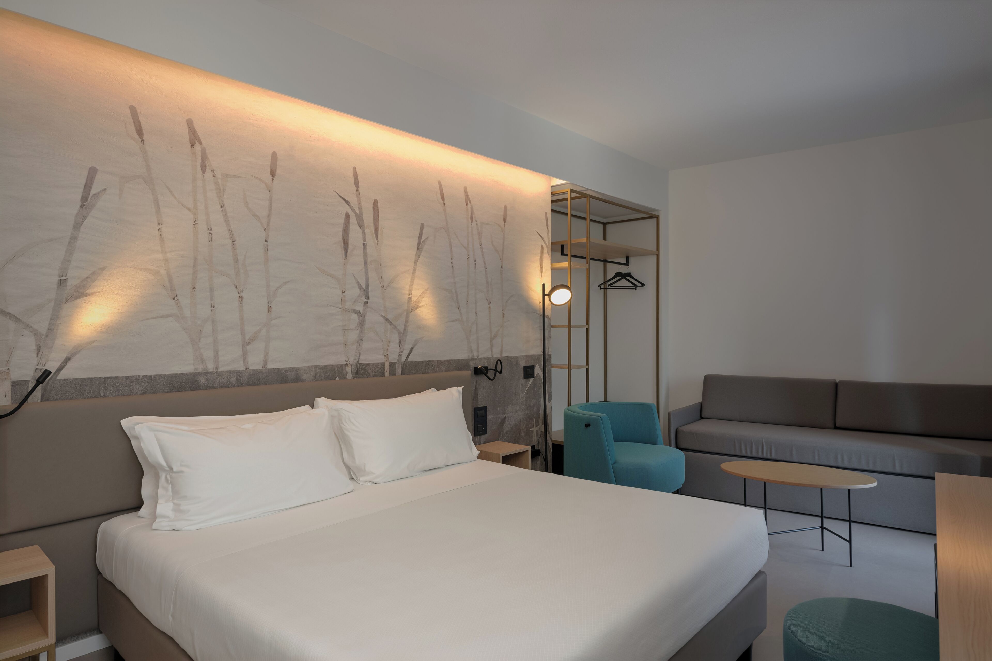 A modern hotel room with a large bed, a sofa, a small table, and a wall-mounted light fixture.