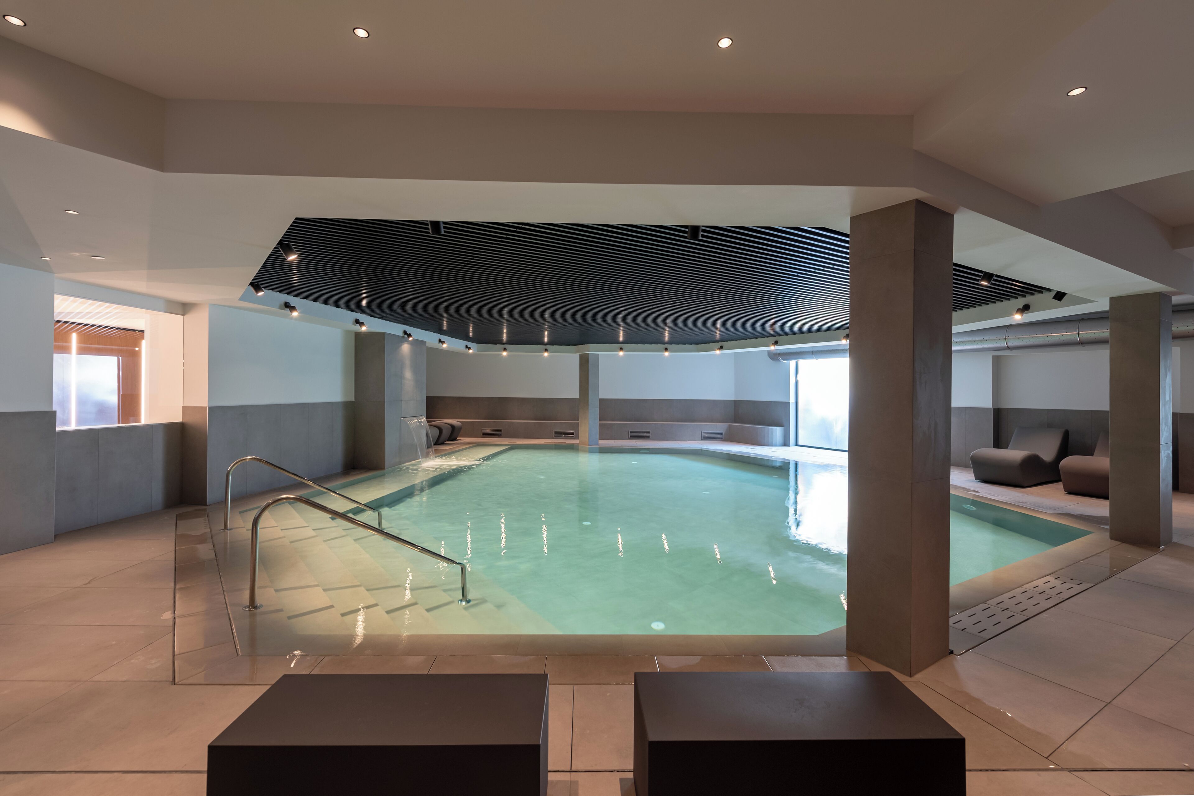 Ein modernes, innenliegendes Schwimmbad im minimalistischen Stil, mit einem zentralen Beckenbereich, Sitzbereichen und einer schrägen Decke mit integrierter Beleuchtung.