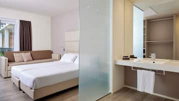 A modern hotel room with a bed, a sofa, a glass partition, a bathroom with a mirror, and a sink.