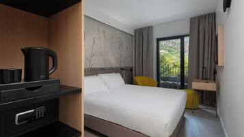 A modern hotel room with a bed, a desk, a chair, a coffee maker, and a window with curtains.
