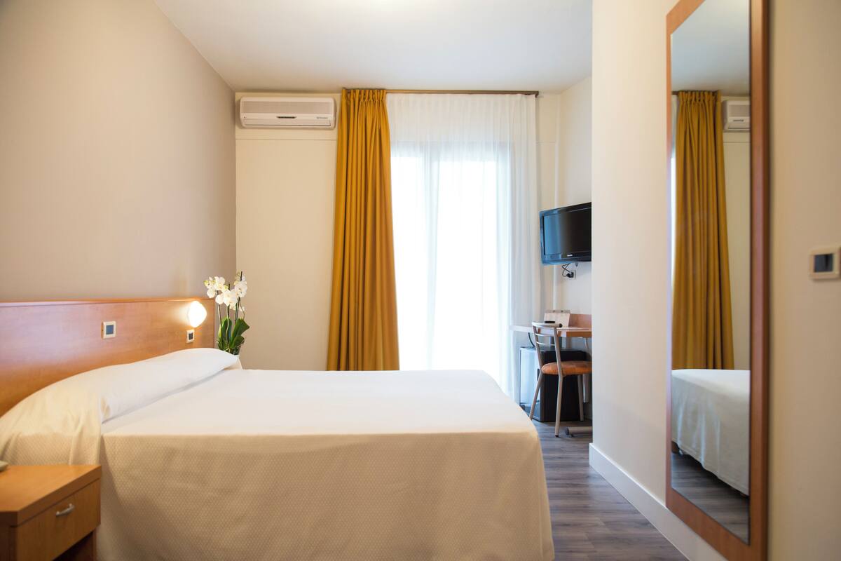 Une chambre d’hôtel avec un lit, une table de chevet et un bureau avec une chaise. Un téléviseur est fixé au mur, et un miroir se trouve sur le mur opposé.