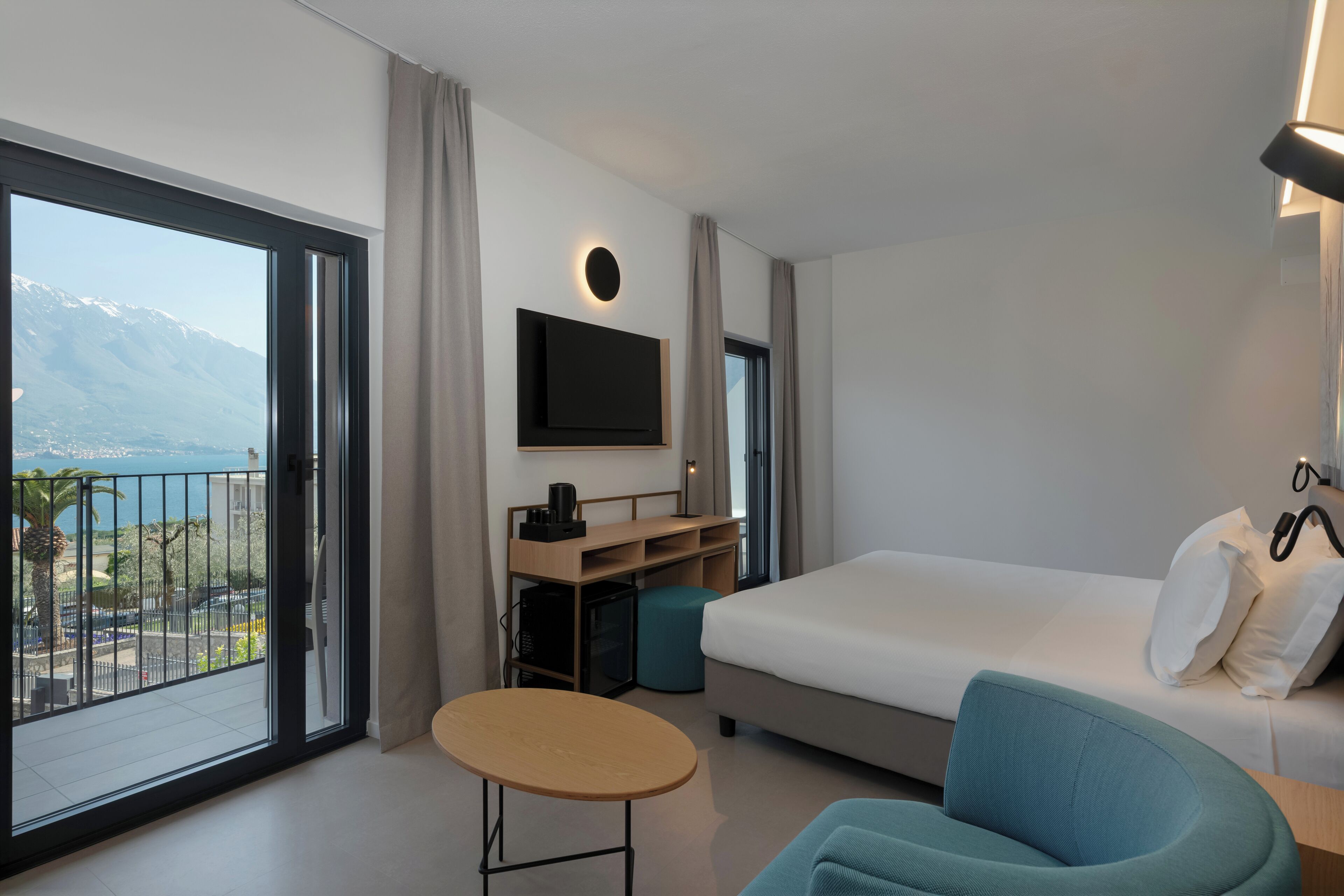 A modern hotel room with a large bed, a desk with a flat-screen TV, a small round table, and a view of a mountain and body of water.