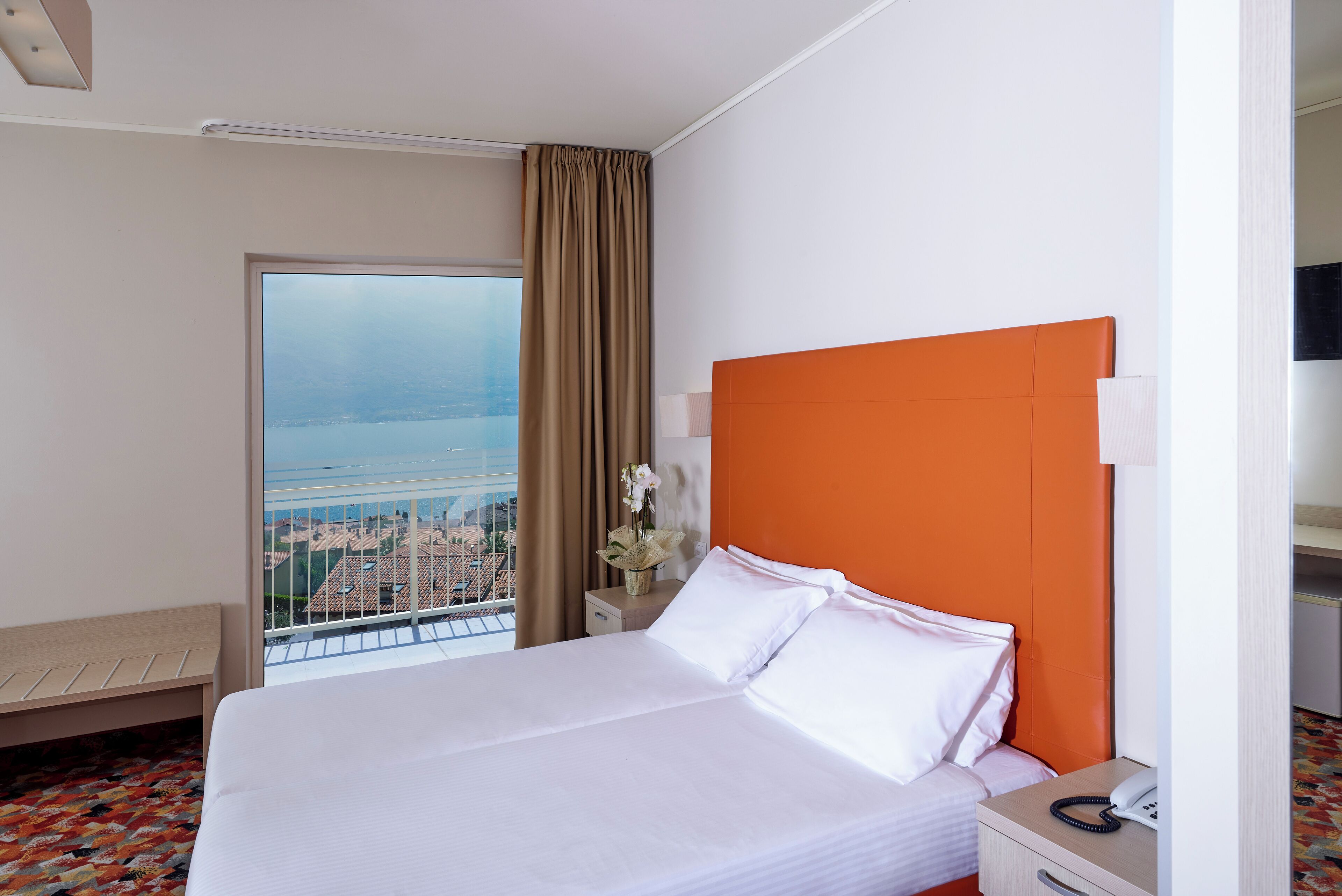 Habitación de hotel con cama, mesita de noche con teléfono, una mesita de noche con un jarrón de flores, una ventana con vista a la ciudad y un banco junto a la ventana.