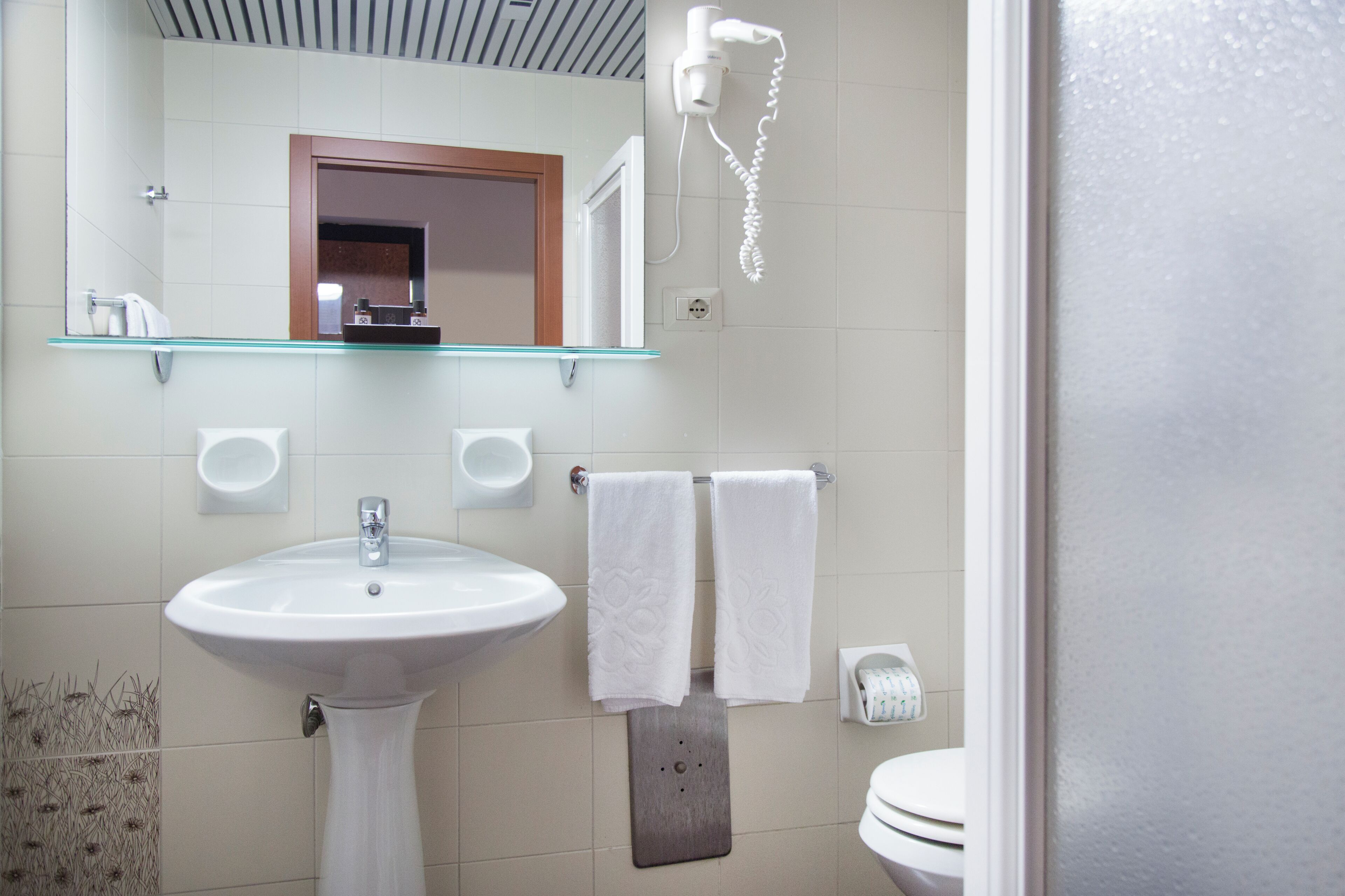 Et badeværelse med et pædebordsvask, to håndklæder hængende på en vaskekurv, en glasshylde over vasken, en hårtørrer monteret på væggen, og et toilet.