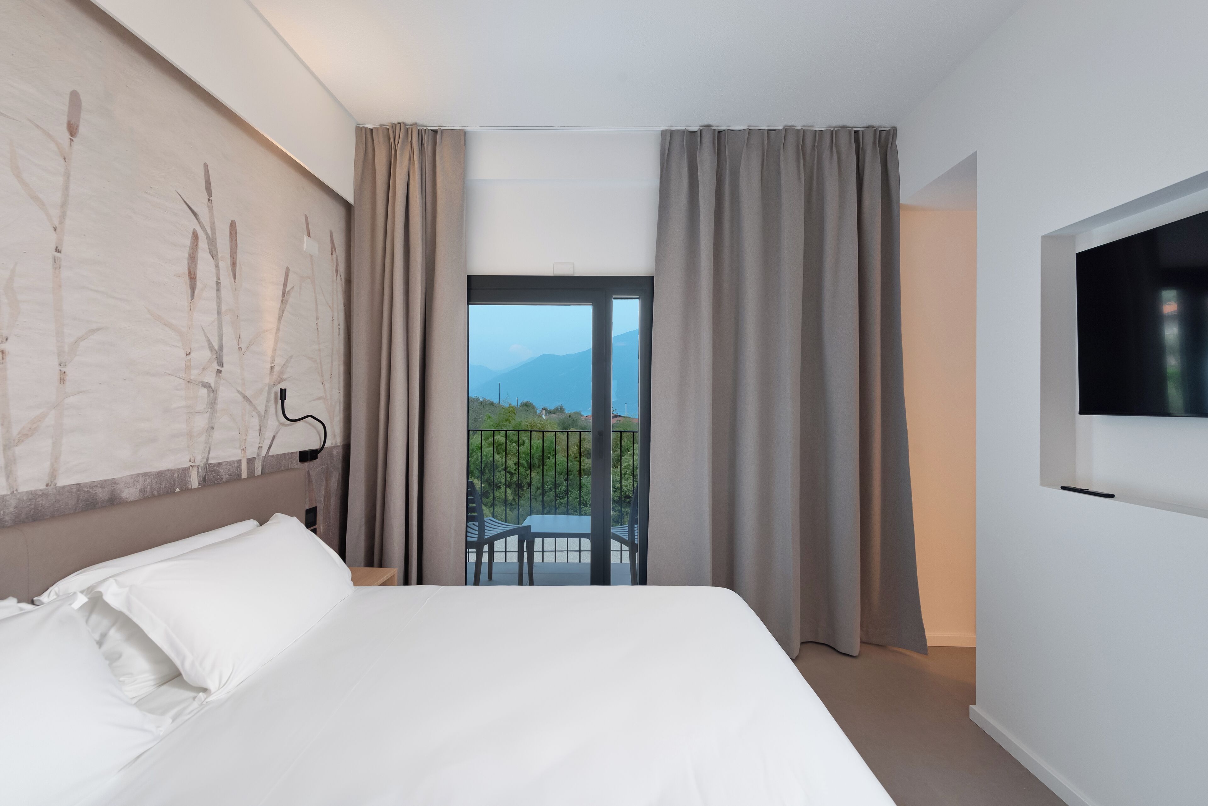 Habitación de hotel con cama, televisión, balcón con vistas a montañas y un cuadro en la pared.