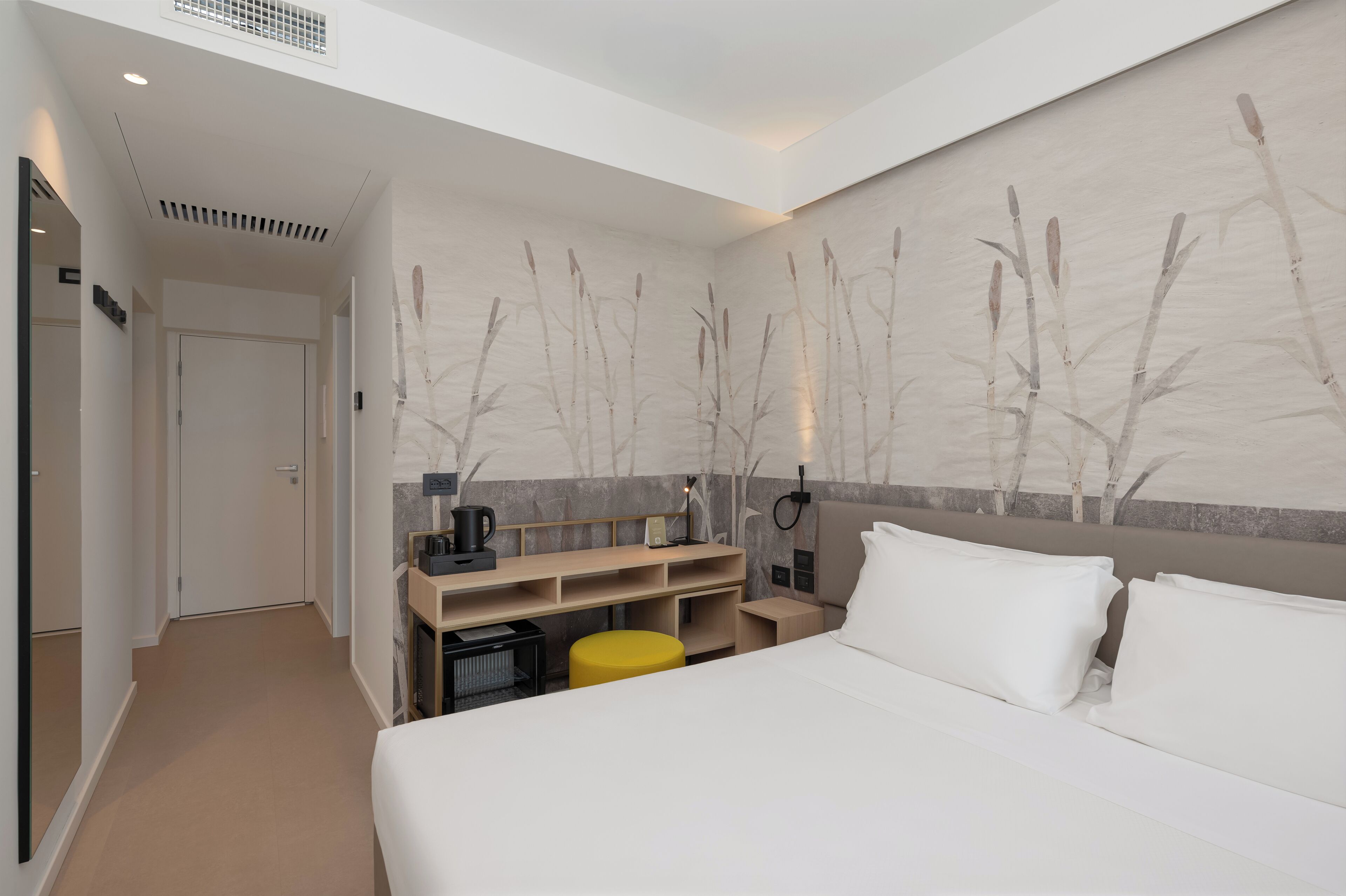 Ein Hotelzimmer mit Bett, Schreibtisch, Stuhl, kleinem Tisch, Nachttisch und einer Wand mit Pflanzenmotiv.