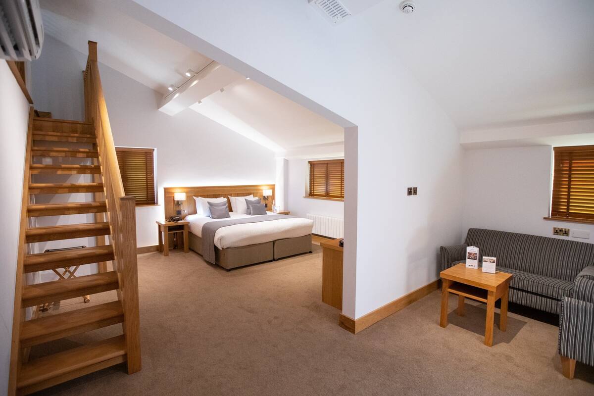 A hotel room with a wooden staircase, a bed with a headboard, a small table, and a sofa.