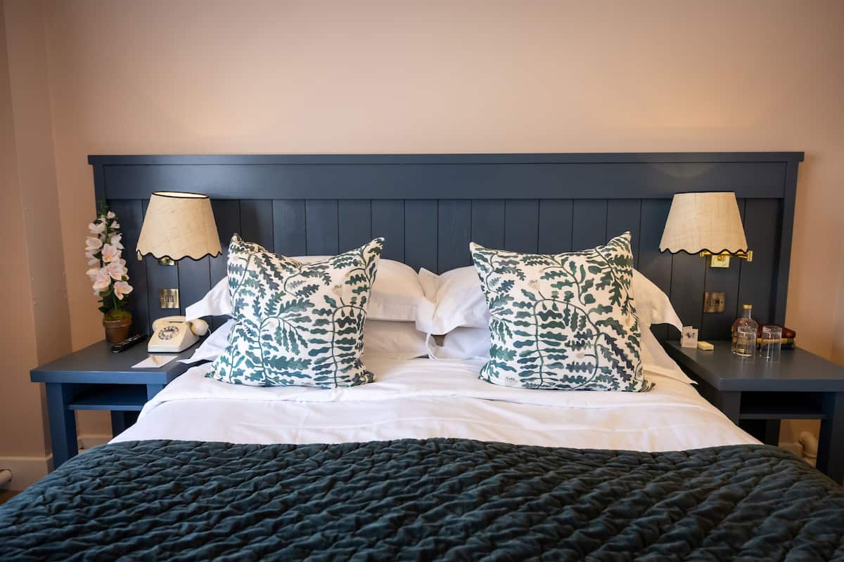 A neatly made bed with a dark quilt, two patterned pillows, and a matching headboard. There are two bedside tables with lamps and a small vase of flowers.