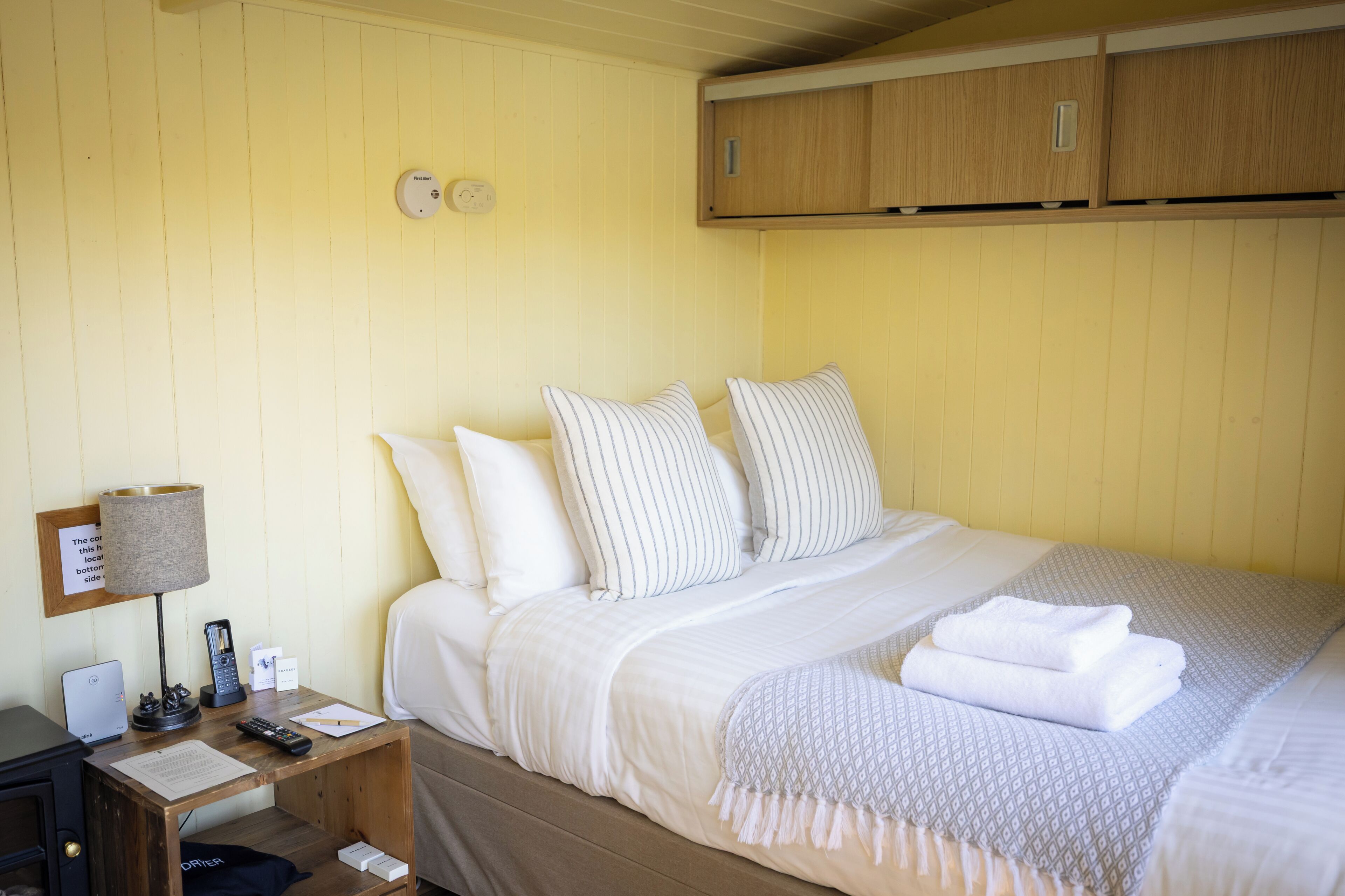 A neatly made bed with white bedding and striped pillows, a bedside table with a lamp, a remote control, and a laptop.