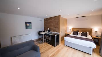 Quarto de hotel moderno com uma cama, sofá, pequena cozinha e parede de tijolos decorativa.