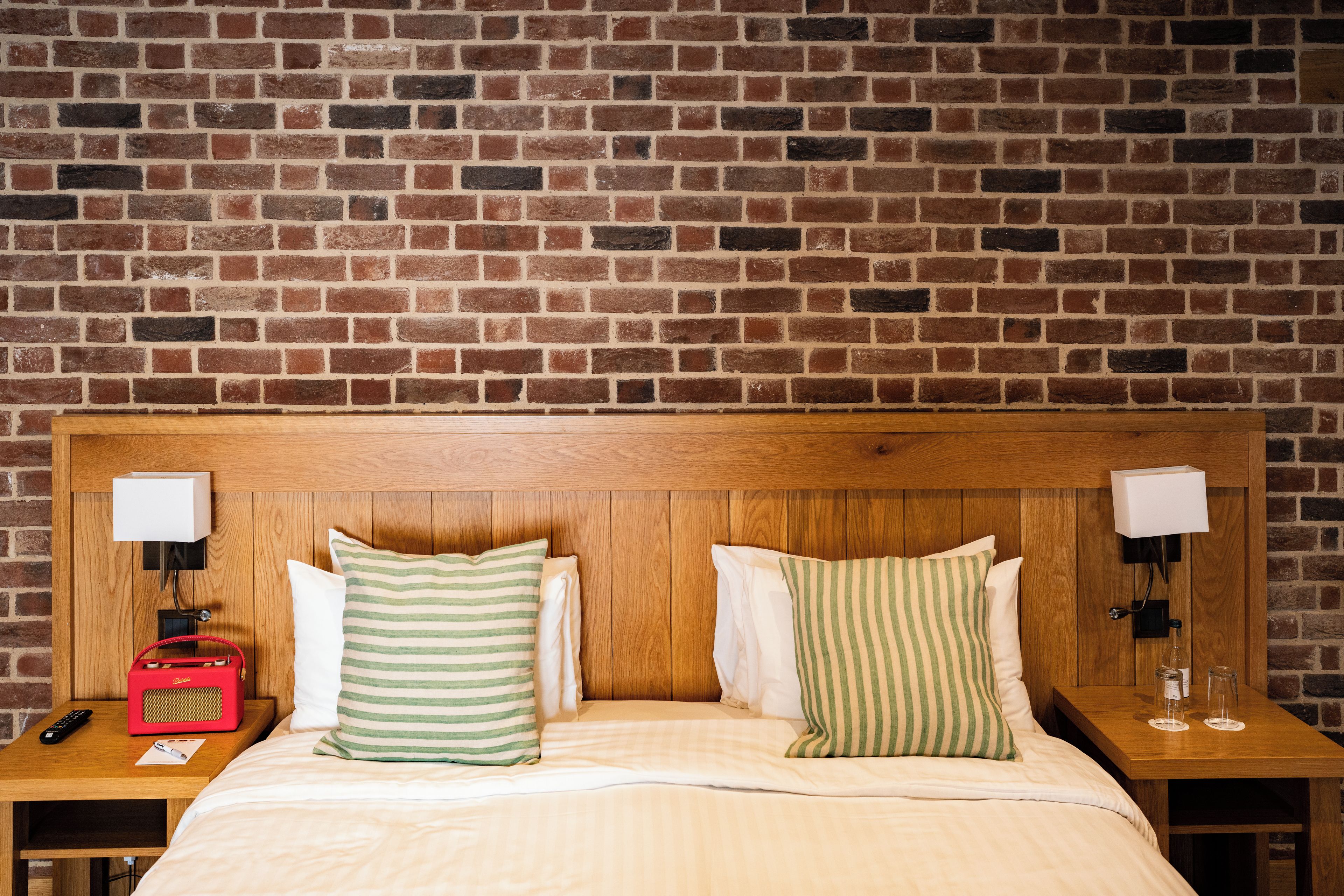 Une chambre d’hôtel avec un mur en briques, une tête de lit en bois, deux tables de chevet, un lit avec une literie blanche et des oreillers à rayures.