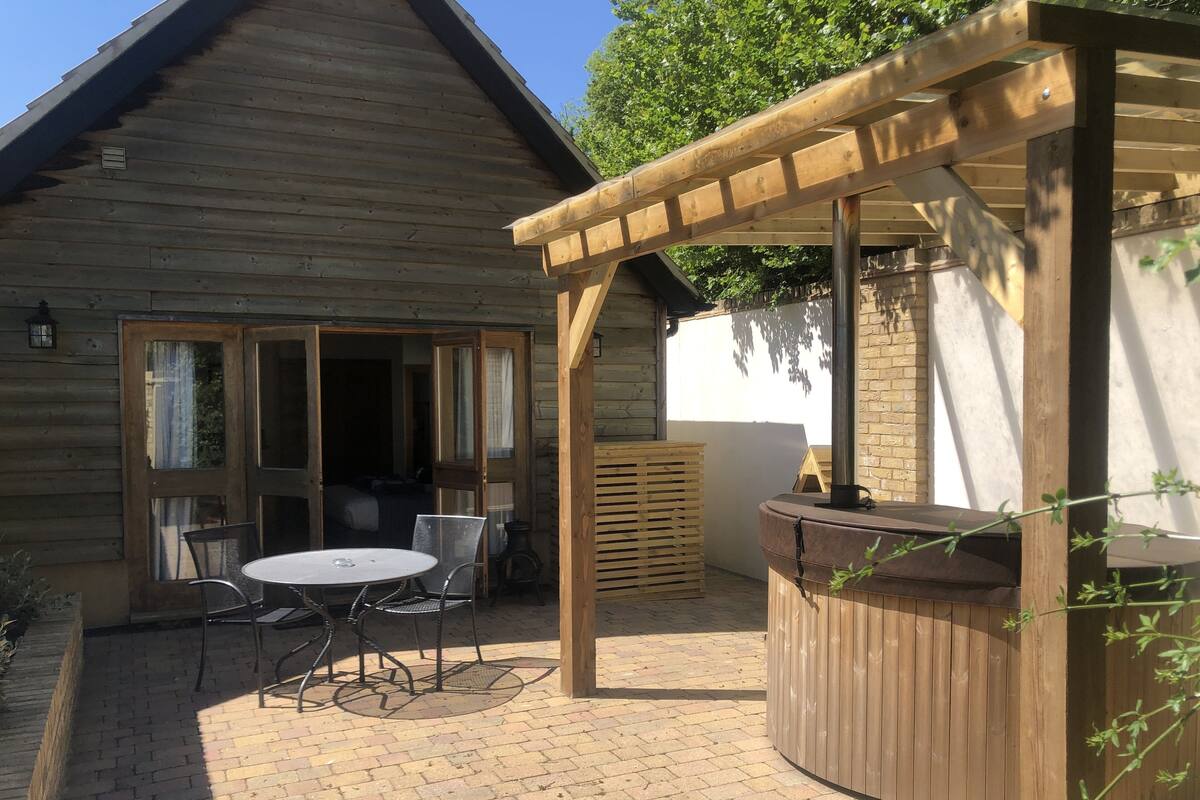 A wooden cabin with a patio area featuring a round table and chairs, a wooden pergola, and a hot tub.