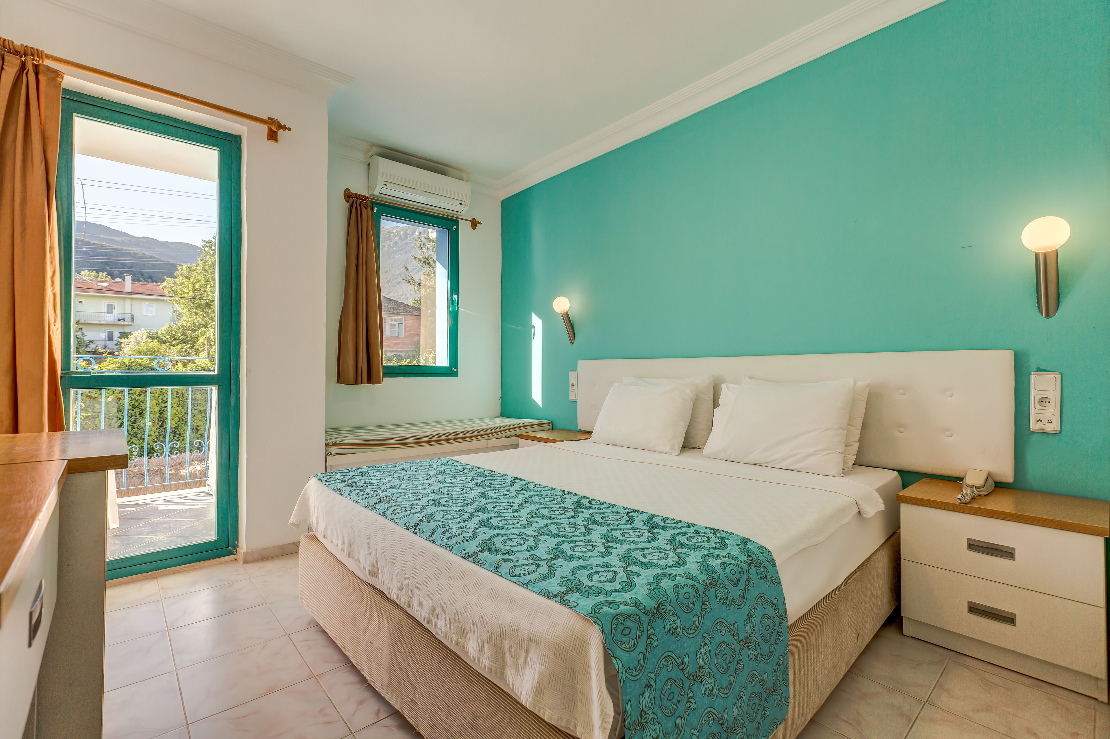 A bedroom with a bed, a nightstand, a window with curtains, and a view of buildings outside.