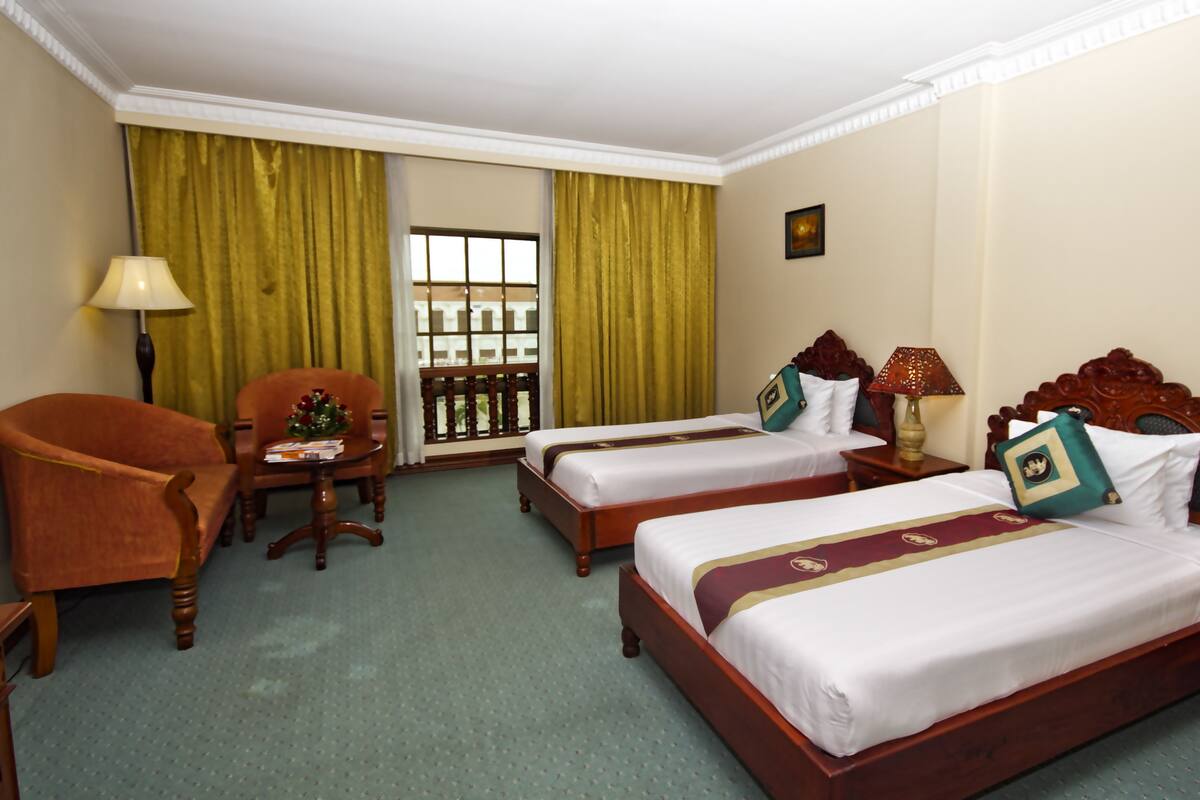 A hotel room with two beds, a desk, a chair, a window with curtains, and a lamp.