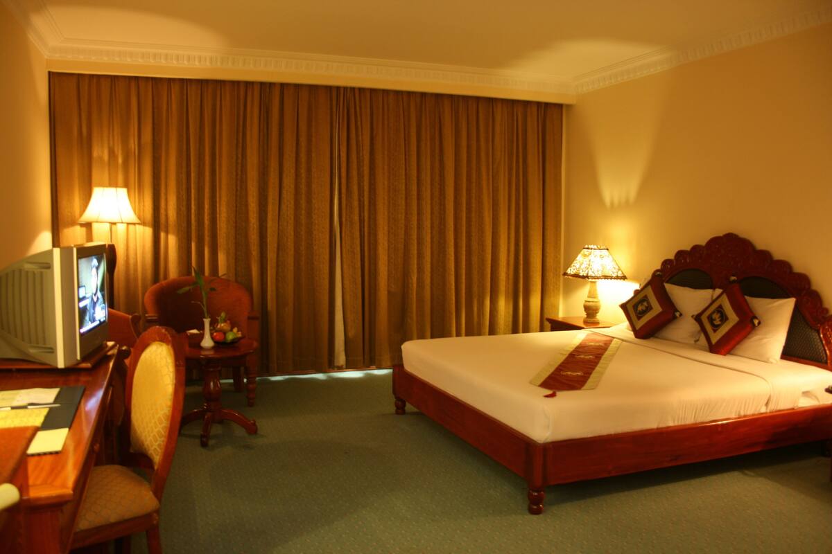 A hotel room with a bed, a desk with a television, a chair, and a lamp.