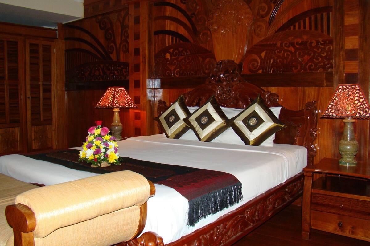 A wooden four-poster bed with ornate carvings, two lamps, and a floral arrangement on the bed.