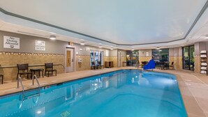 Una piscina interior con un cartel de prohibición de saltos, un cartel indicando presencia de socorrista, y una zona junto a la piscina con sillas y un área de descanso cubierta.