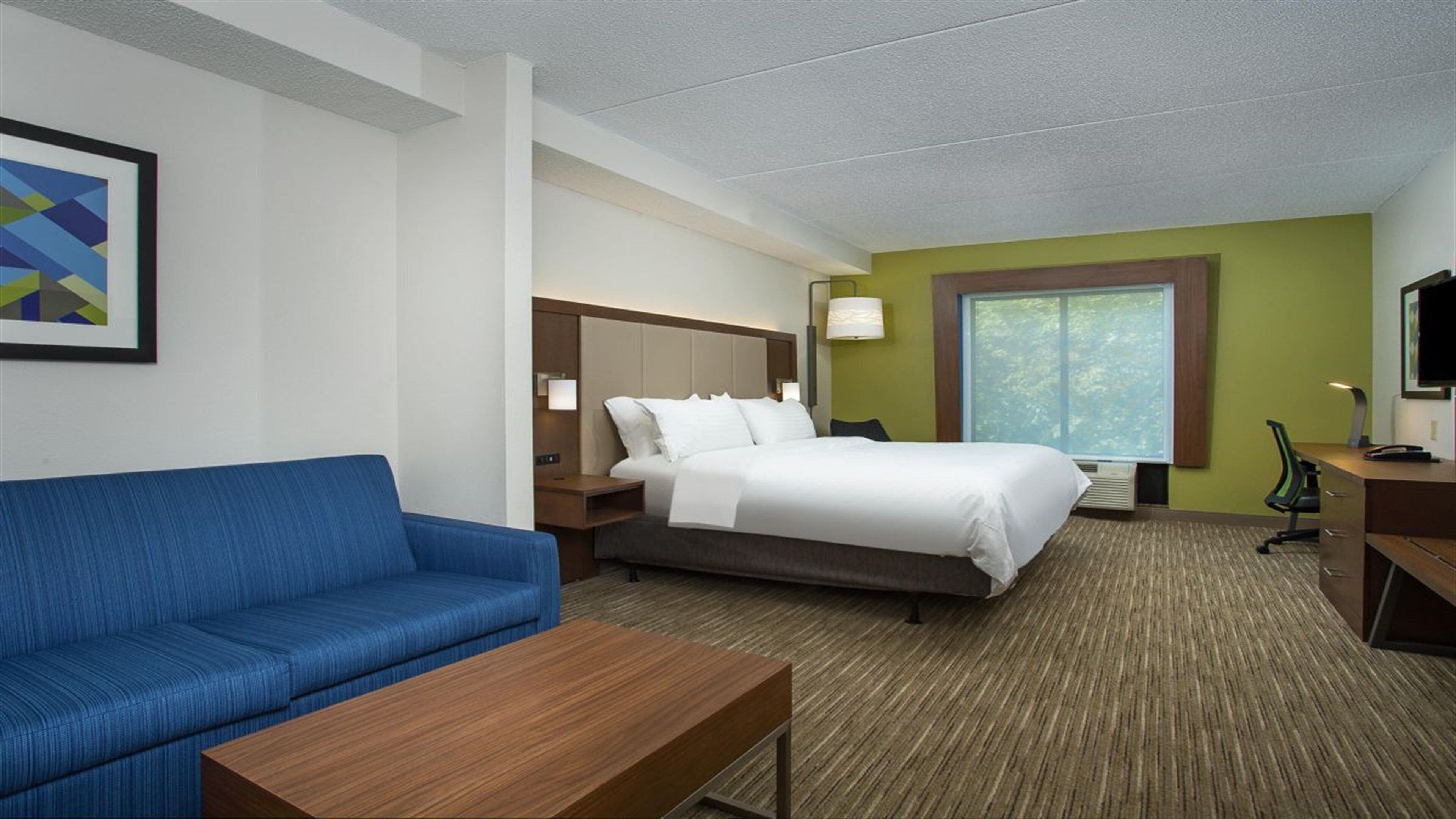 A hotel room with a bed, a blue sofa, a wooden table, a desk, a lamp, and a window.