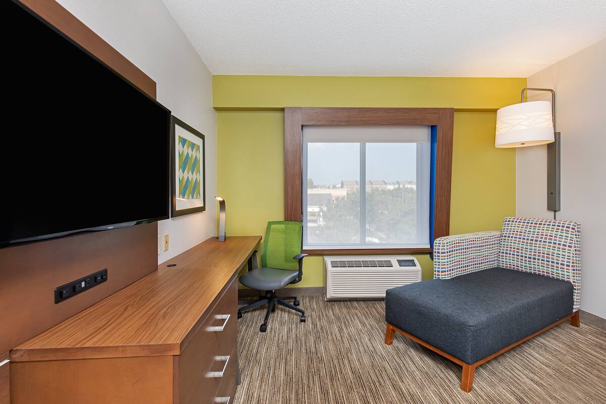 A hotel room with a desk, chair, bed, window, and TV.
