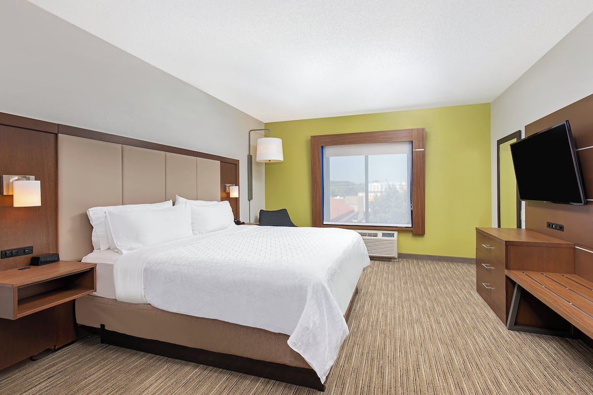 A hotel room with a large bed, a desk, a TV, and a window with a city view.
