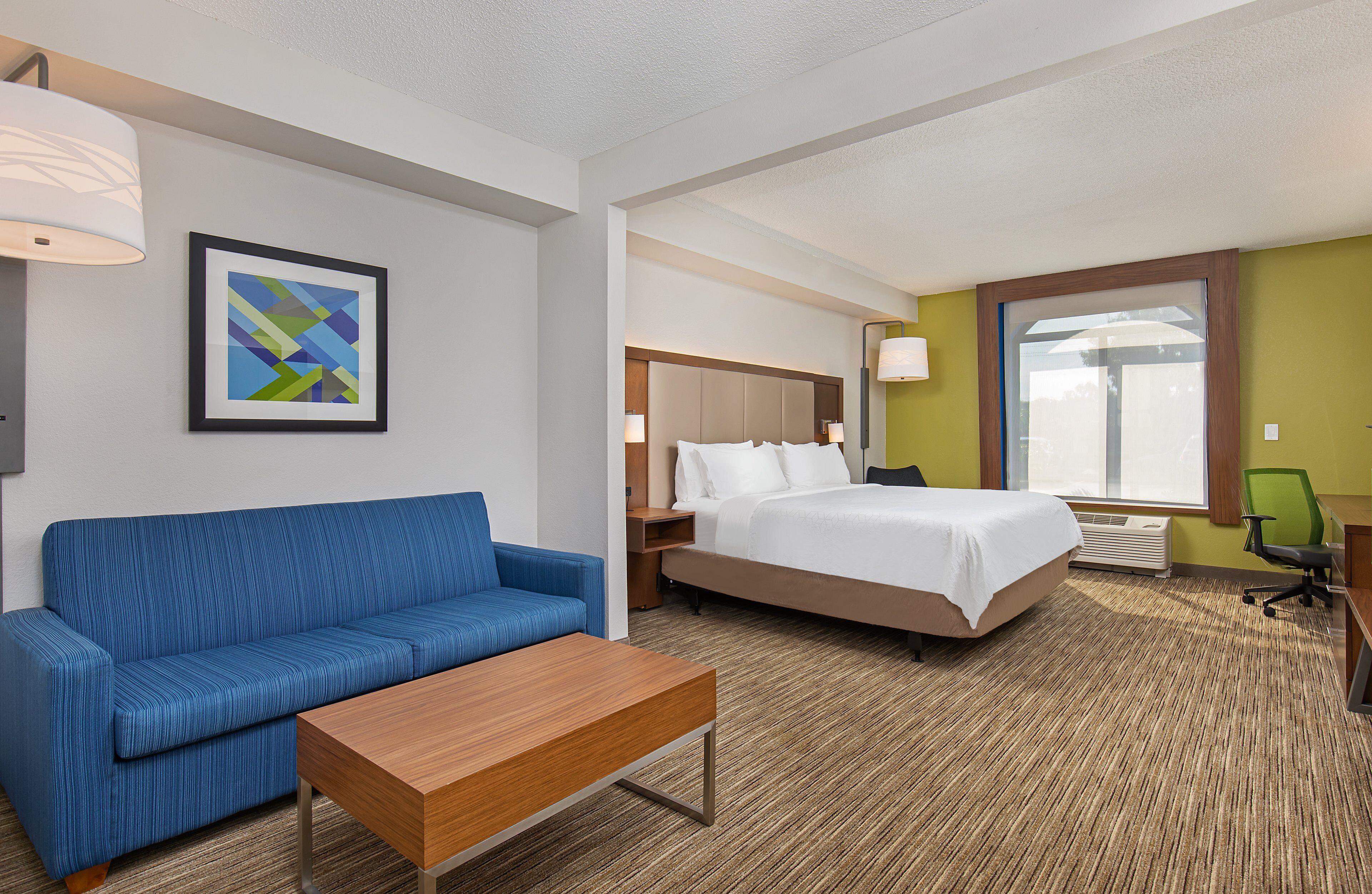 Habitación de hotel con cama, sofá azul, mesa de centro de madera, un cuadro en la pared y ventana con vistas.