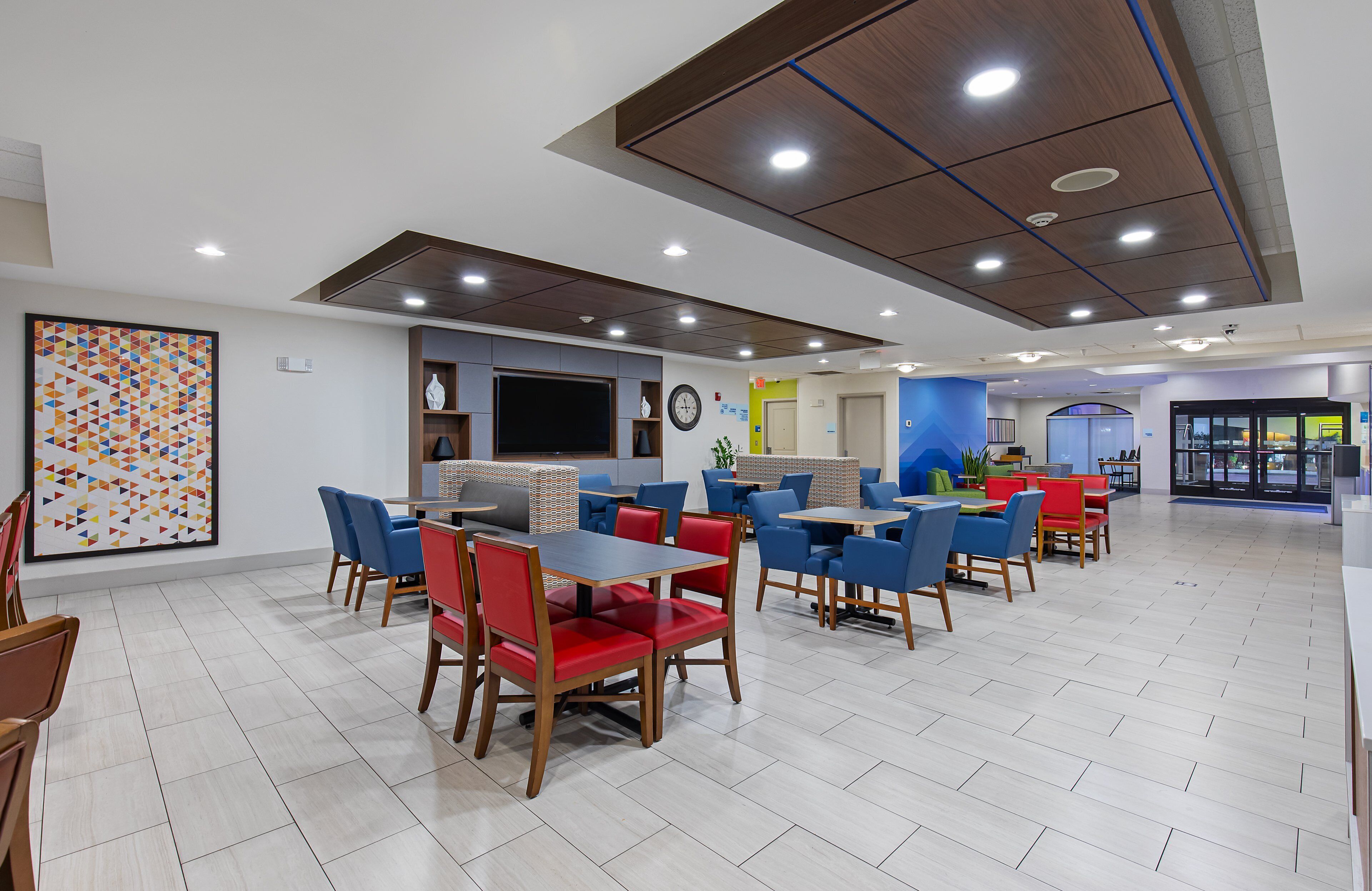 A modern dining area with multiple tables and chairs, a large clock, and a television on the wall.