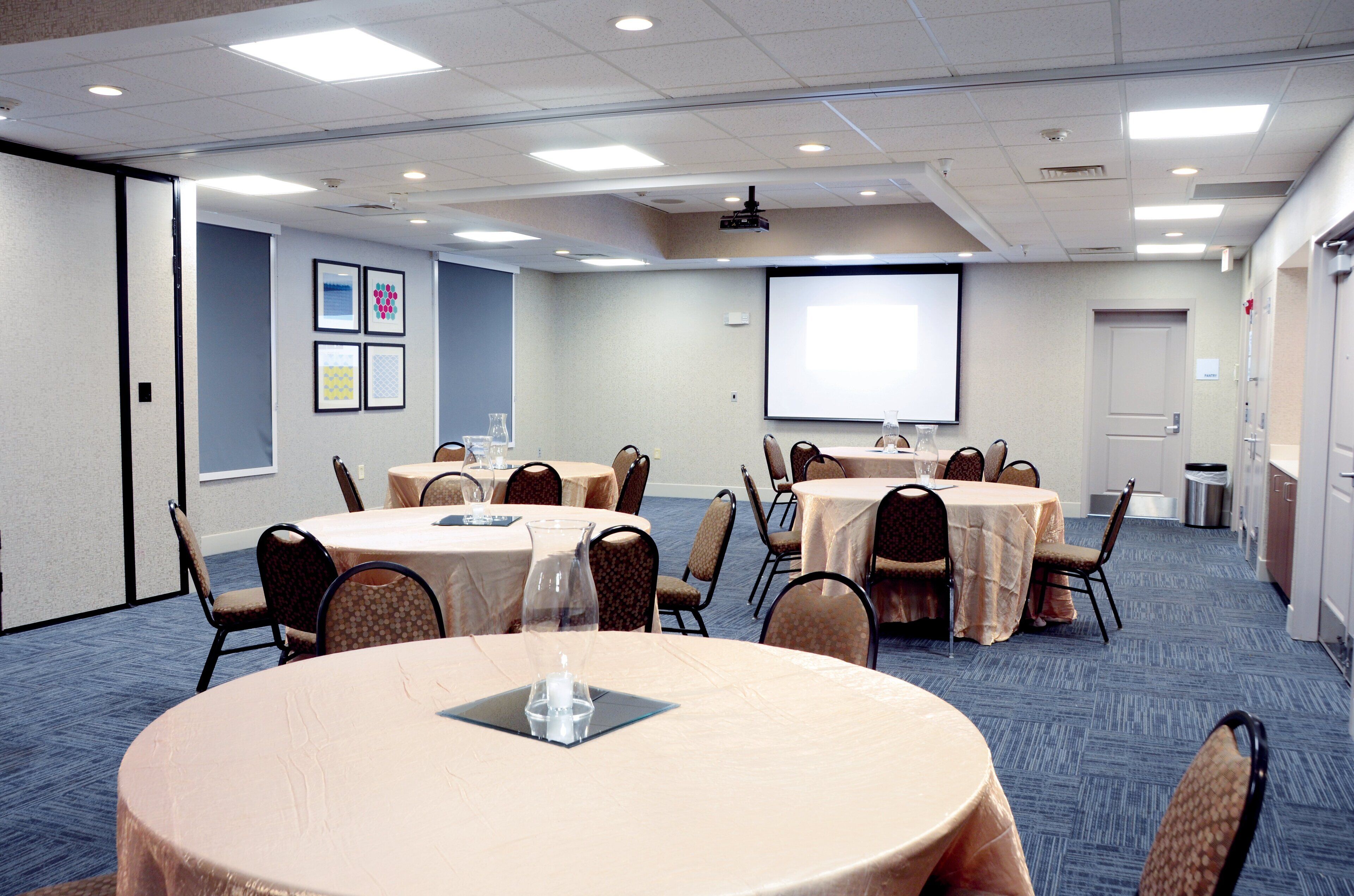 A conference room with multiple tables and chairs, a projector screen, and framed pictures on the wall.
