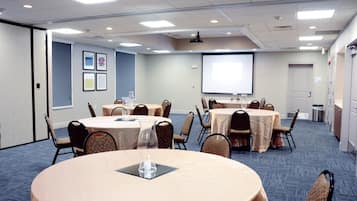 A conference room with multiple tables and chairs, a projector screen, and framed pictures on the wall.