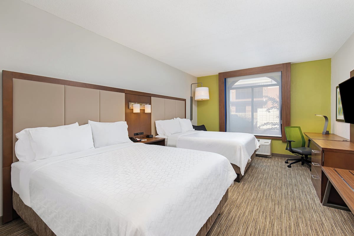 A hotel room with two beds, a desk, a chair, a nightstand, and a window.