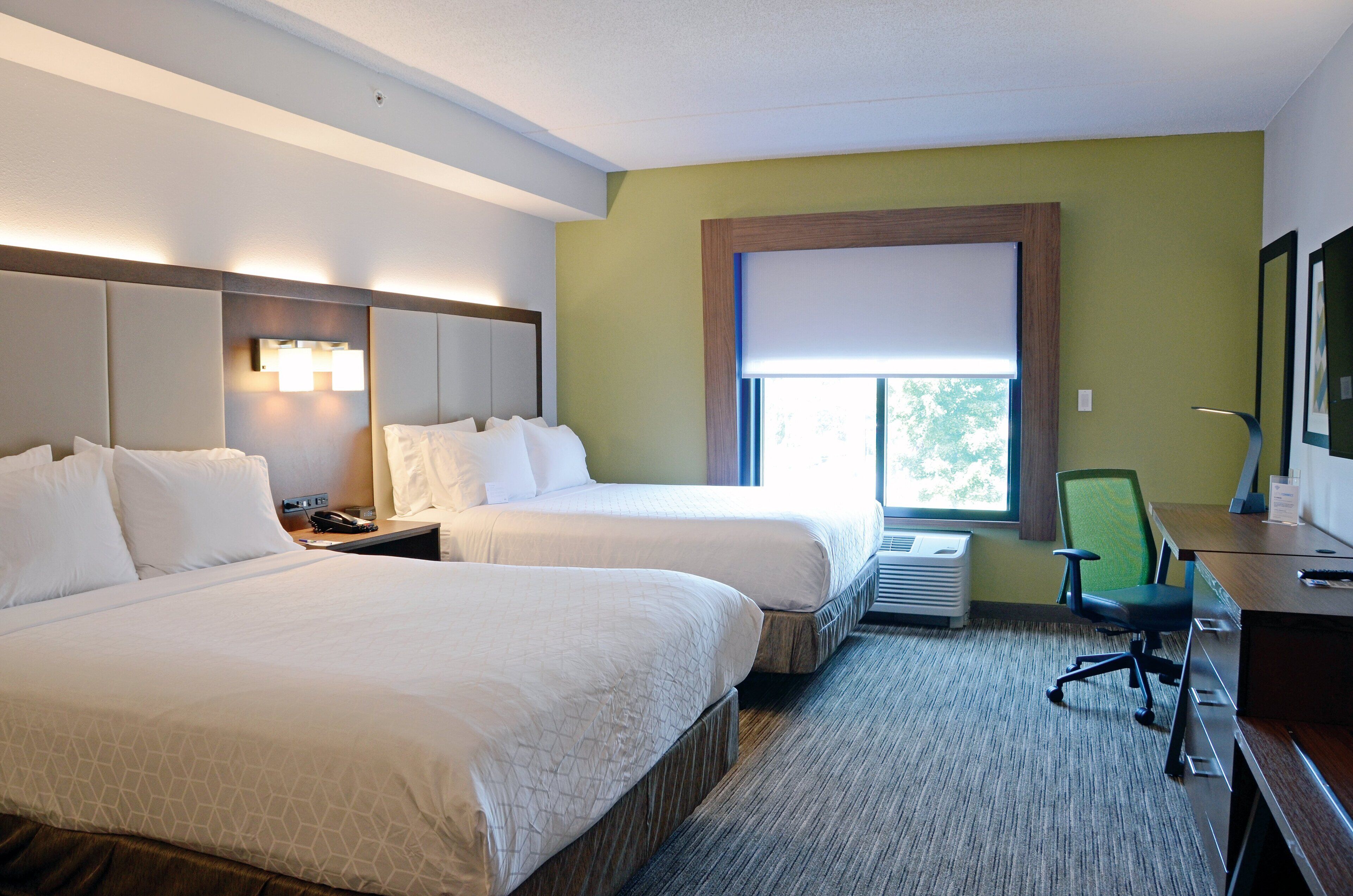 A hotel room with two beds, a desk with a chair, a window with a view, and a wall-mounted air conditioning unit.