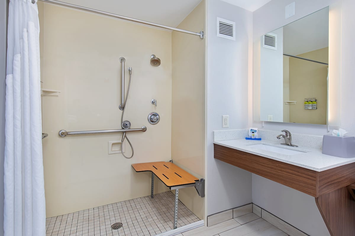 A shower with a grab bar, a small wooden bench, and a white sink with a mirror above it.