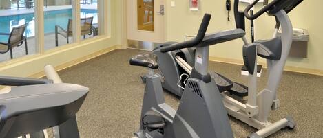Un gimnasio con cintas de correr y elípticas, una piscina visible a través de puertas de cristal, y un cartel que detalla las instalaciones del centro.