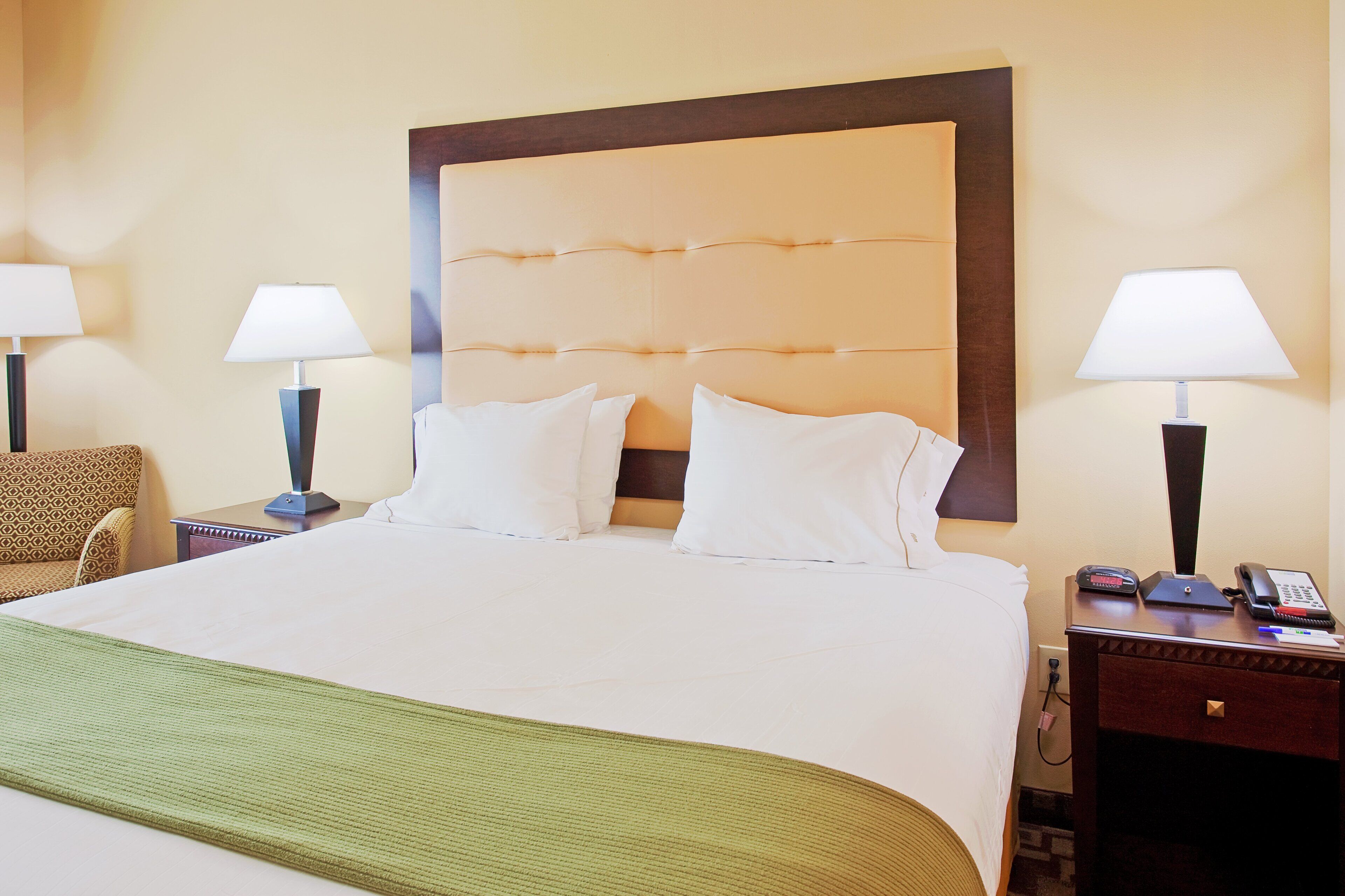 Habitación de hotel con una cama grande, dos lámparas de noche, una silla, una mesita de noche con un teléfono y una manta verde.