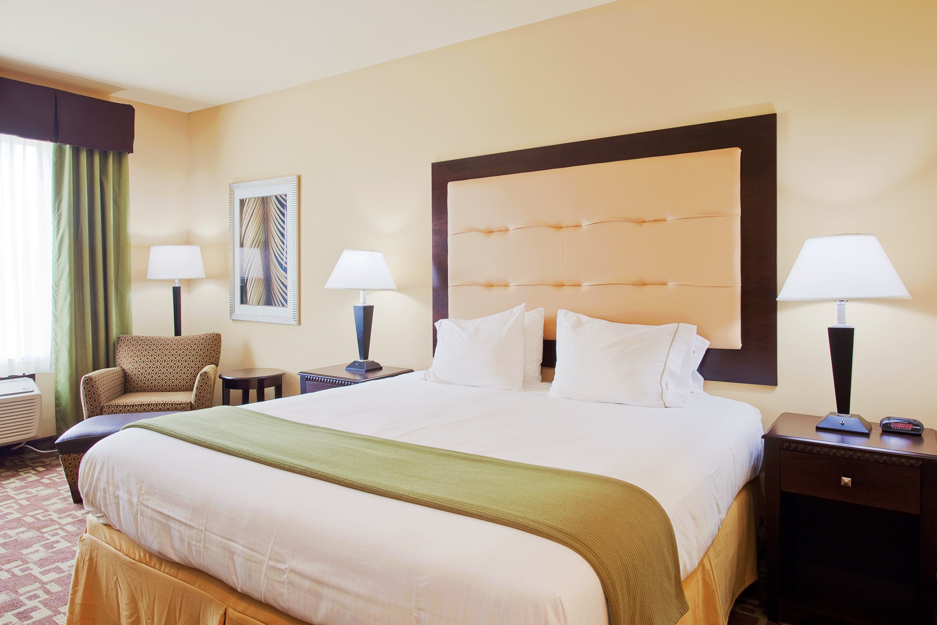 Une chambre d’hôtel avec un grand lit, deux tables de chevet avec des lampes, une chaise, une œuvre d’art accrochée au mur et une fenêtre avec des rideaux.